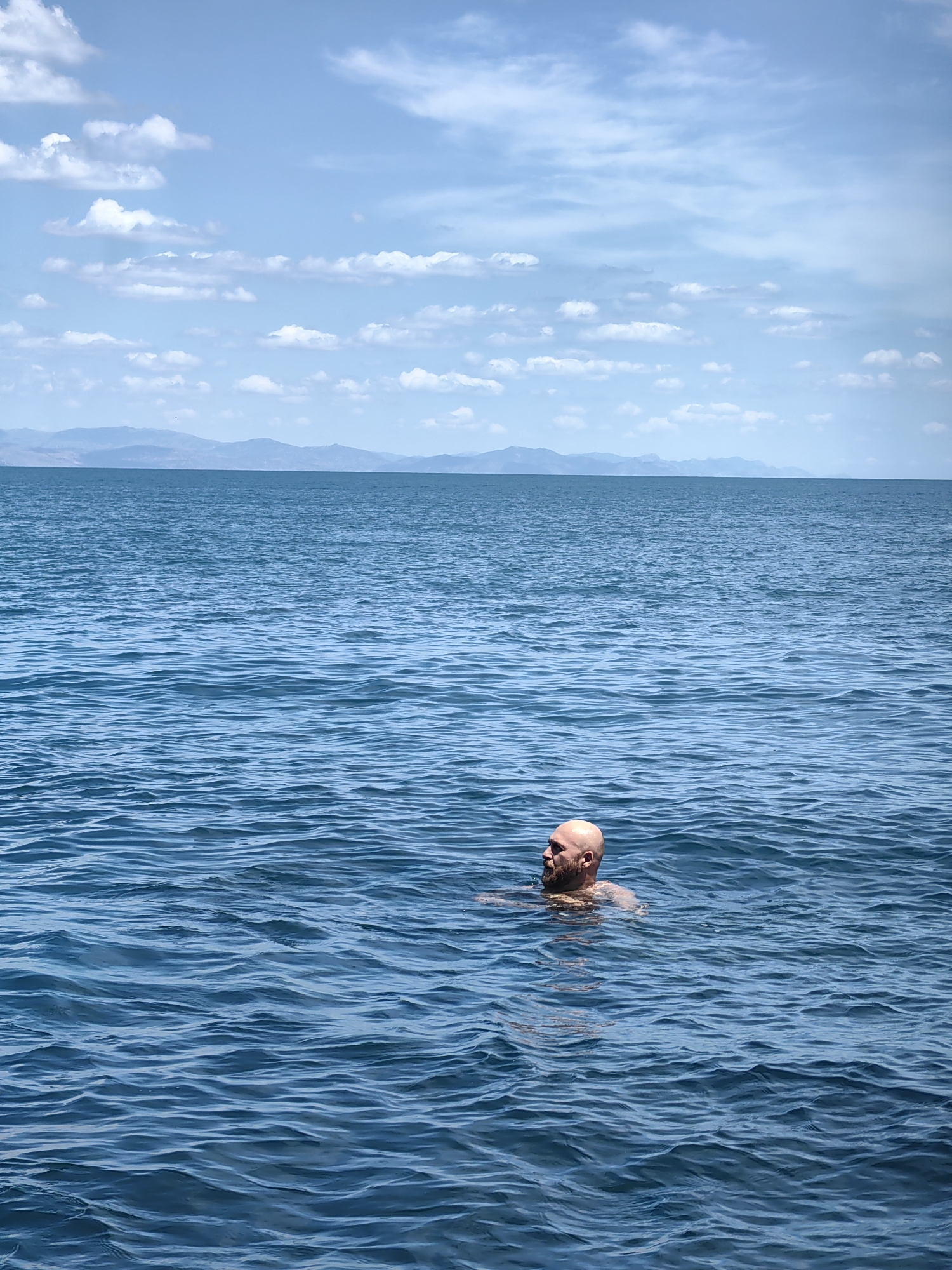 In the city of Utios, Ukraine, a bald, bearded man is swimming in the calm, blue waters of a large body of water. The man is positioned in the lower right quadrant of the frame, with his head and shoulders visible above the surface. The water is slightly rippled, reflecting the bright, partly cloudy sky above. In the distance, a hazy, mountainous coastline can be seen along the horizon. The overall scene depicts a serene and tranquil outdoor environment.
