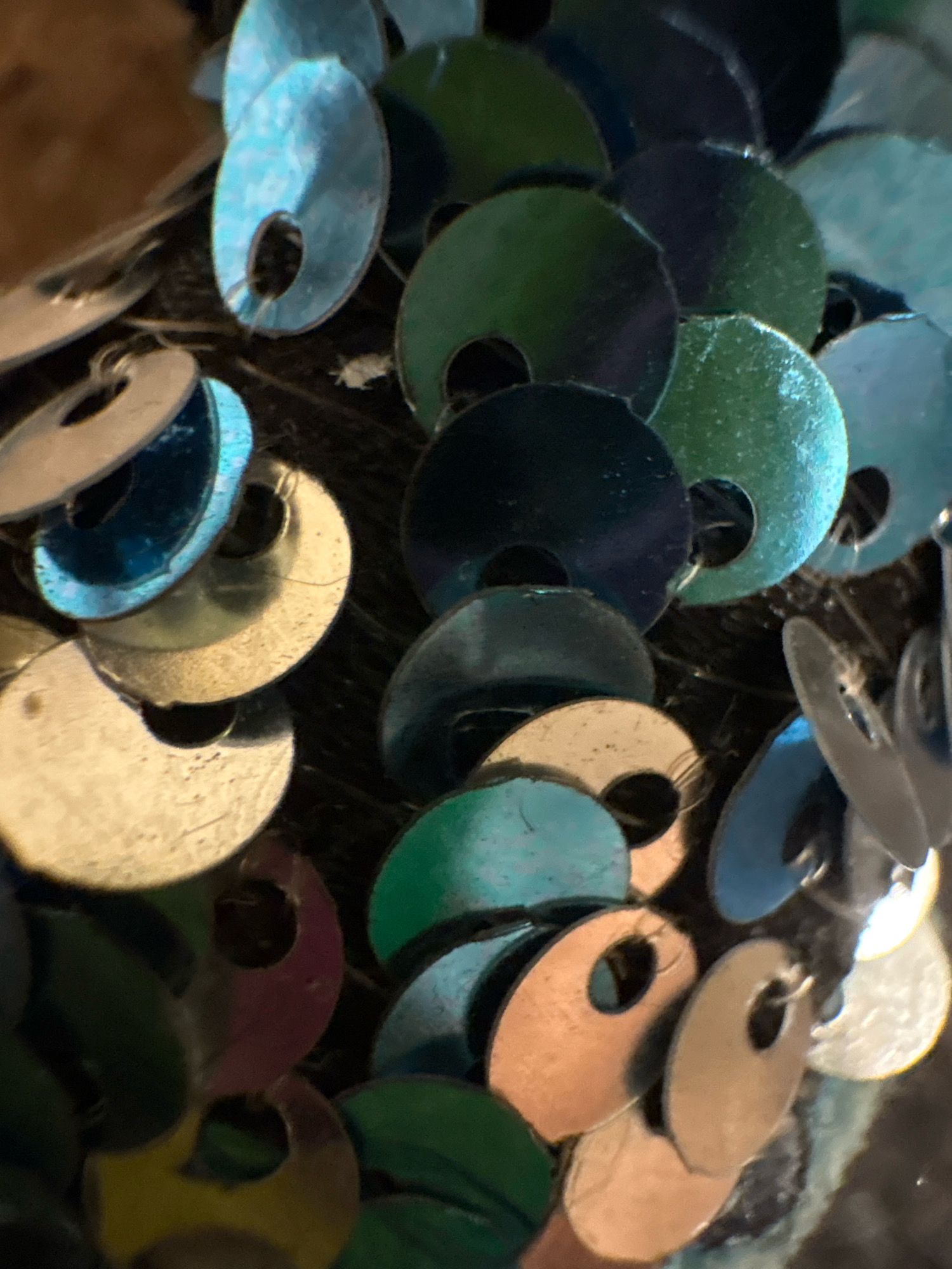 The image is a close-up, macro shot of a collection of iridescent, flat, circular sequins. The sequins are predominantly in shades of blue, green, and silver, with some appearing to have a golden or copper hue. They are overlapping and densely packed, suggesting they are part of a larger fabric or craft project. Each sequin has a small hole in the center, indicating they are likely sewn or strung together. The lighting creates strong highlights and reflections on the metallic surfaces of the sequins, giving them a shimmering appearance. The background is dark and out of focus, providing no discernible environmental context other than that the sequins are likely arranged on a dark surface or material. There are no people or actions depicted in the image, and no elements to suggest a specific location in Ashvem, India.