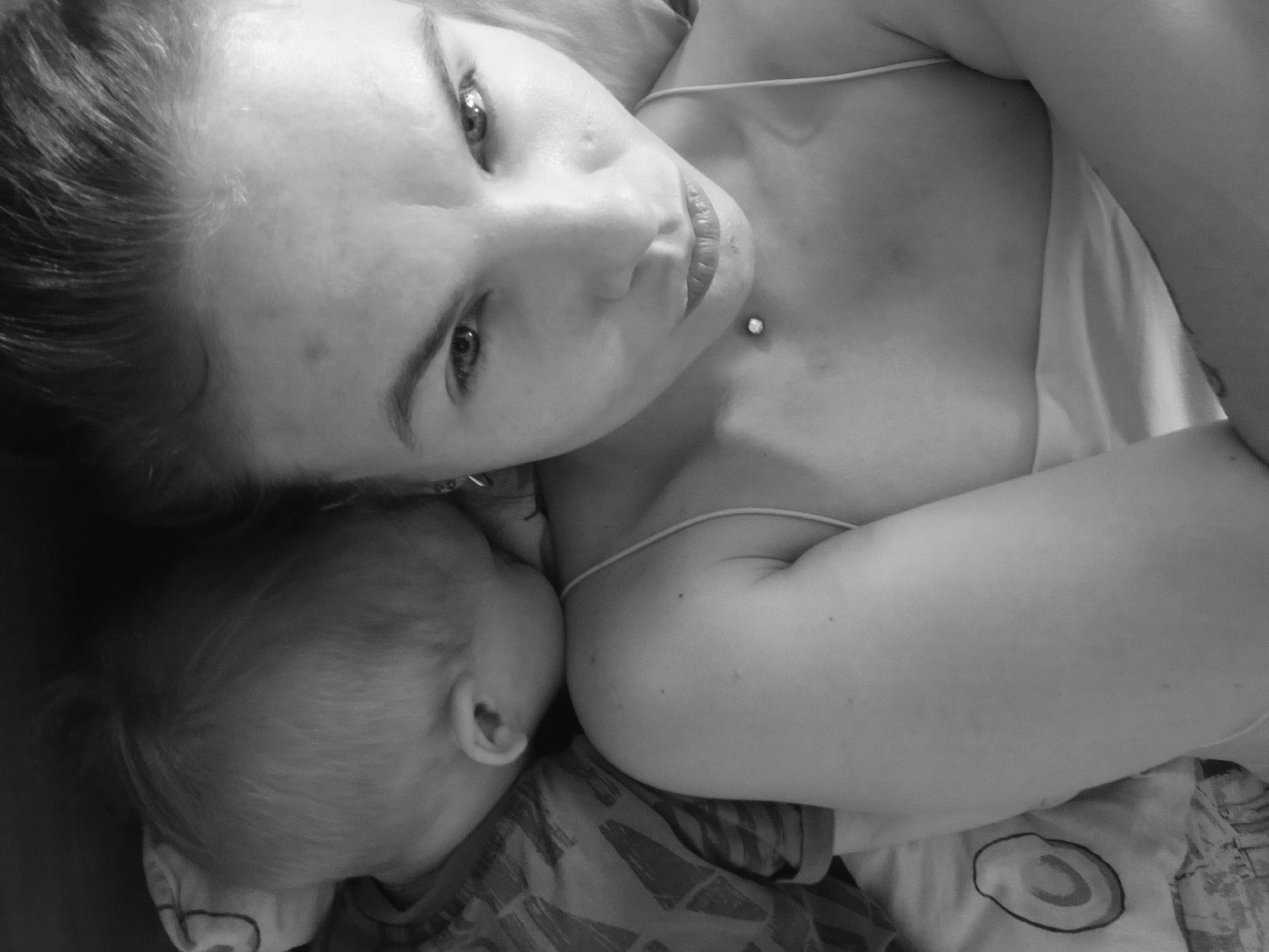 A black and white, high-angle shot depicts a fair-skinned woman lying down with a baby. The woman's face is visible, with eyes looking towards the camera, and a sternoclavicular dermal piercing is visible. The baby is positioned to the left, with its head near the woman's face. The woman is wearing a spaghetti-strap top. The baby is clothed in a patterned top. The background is unclear. There is no information available to determine if this image was taken in Chaikovsky, Russia.