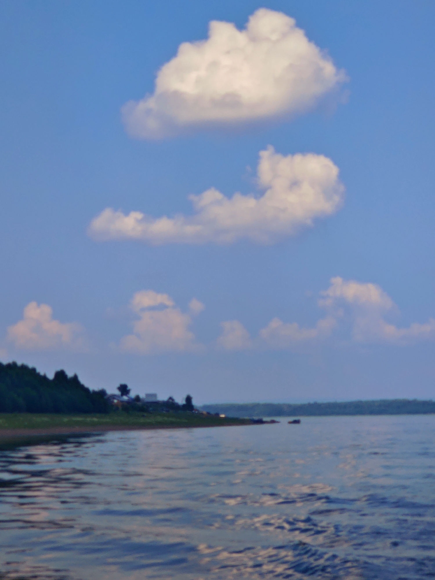 A calm body of water is in the foreground of the image, with a shoreline to the left featuring a line of trees and some distant buildings. The sky above is blue and clear, with a few scattered white clouds. The image is taken from a low angle, suggesting the vantage point is from a boat or the water's edge. The overall scene is tranquil and depicts a natural landscape. There are no people or animals visible in the image. The location in the city of Погодаево, Russia, is not explicitly indicated within the visual elements of the photograph itself.