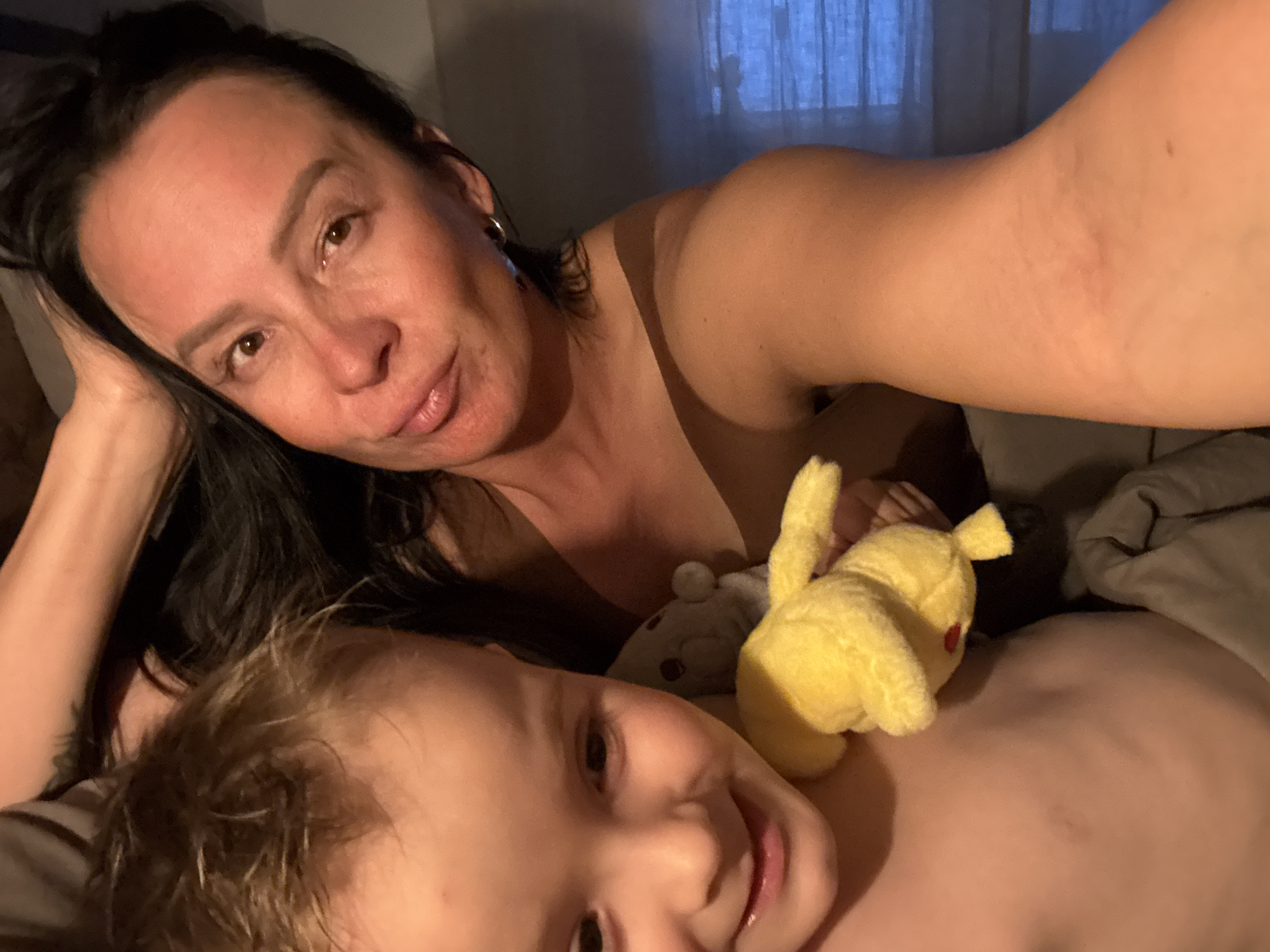 The image is a selfie taken indoors, possibly in a bedroom. A woman with dark hair, wearing a hoop earring, is lying down, with a young child beside her. The child has light-colored hair and is lying on his back. A yellow Pikachu plush toy rests on the child's chest. A gray stuffed animal is nearby. The background shows a window with curtains. There is no information in the image to determine the location is in Chaikovsky, Russia.