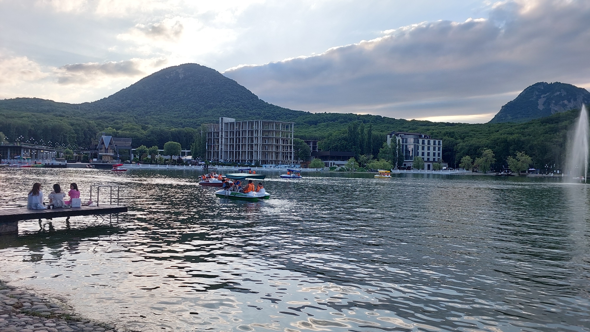 The image depicts a serene lake in the city of Zheleznovodsk, Russia, with a backdrop of lush green mountains under a cloudy sky. In the foreground, three young women are seated on a wooden pier, facing away from the camera, engrossed in conversation or enjoying the view. The lake is populated with several paddle boats, some occupied by groups of people wearing life vests, enjoying a leisurely ride. On the far side of the lake, modern buildings and trees line the shore, with a prominent fountain creating a tall jet of water on the right side of the frame. The overall scene conveys a peaceful and recreational atmosphere.