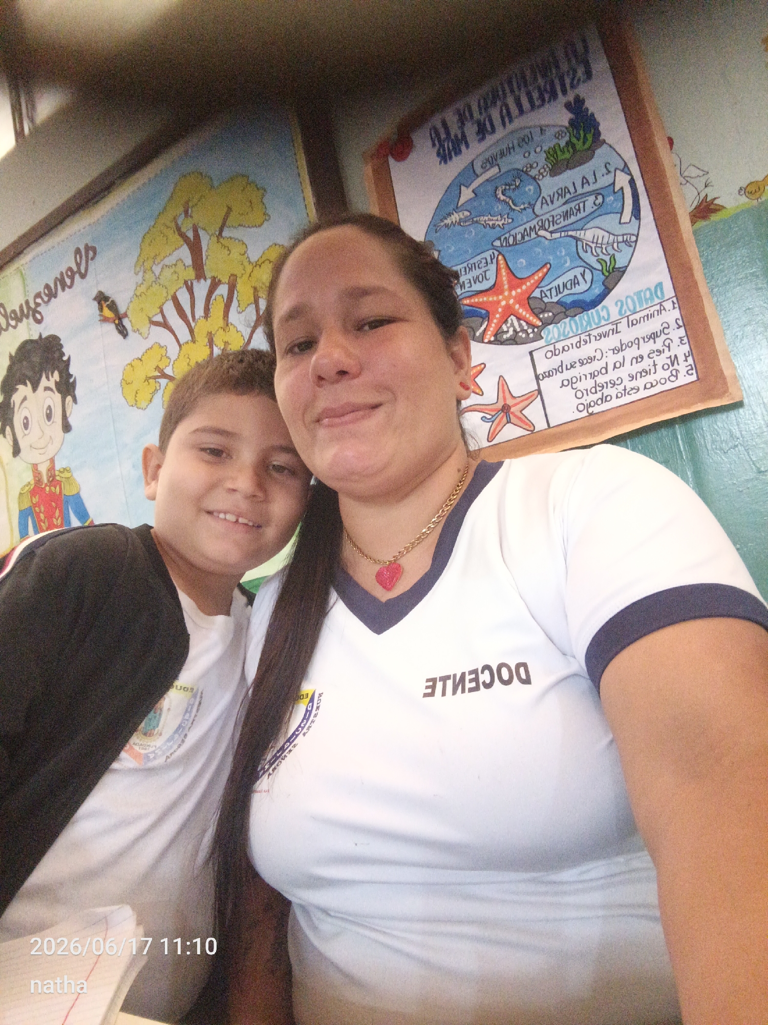 The image shows a selfie taken by a woman and a young boy, likely in a classroom setting. The woman is in the foreground, smiling and looking towards the camera. She is wearing a white t-shirt with dark trim and a red heart-shaped pendant on a gold chain. The boy, positioned to her left, is also smiling. He is wearing a white t-shirt and a black jacket. Behind them, there are colorful posters on the wall. One poster on the left depicts a cartoon character and a tree with yellow leaves. Another poster on the right features a diagram of marine life and text in Spanish. The overall environment suggests an educational or school-related context. The lower left corner of the image contains a timestamp "2026/06/17 11:10" and the word "natha". Based on the provided context, the location is the city of San Sebastián, Venezuela.