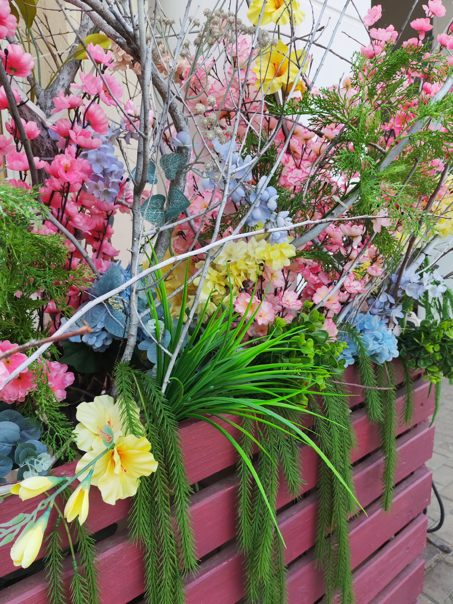 This media file captures a close-up view of a vibrant, artificial floral arrangement housed in a reddish-purple wooden planter. The planter itself is constructed from horizontal slats, creating a layered effect. The arrangement is densely packed with a variety of faux flowers and foliage in shades of pink, yellow, and blue, interspersed with delicate, bare white branches. Green, grass-like artificial plants cascade over the sides of the planter, adding a trailing effect. Amongst the more delicate blooms, a prominent yellow hibiscus-like flower with multiple buds stands out on the left side of the frame, near the bottom. The background is a neutral, light-colored wall, suggesting an outdoor setting, possibly a garden or patio. The lighting appears to be natural daylight, casting soft shadows and highlighting the colors of the artificial flora. No people or text are visible in the image. The context provided indicates this scene is located in Kurgan, Russia.