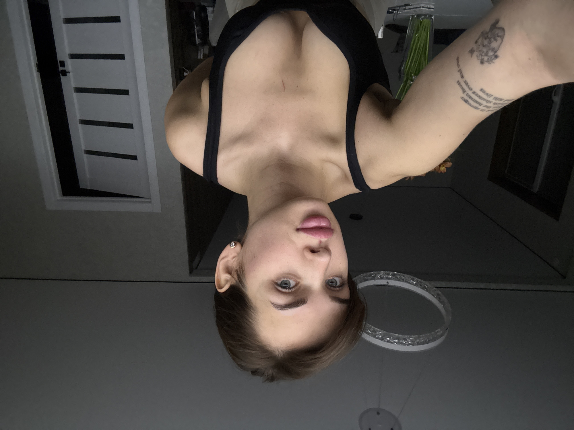 A woman with gray-blue eyes, light brown hair, and fair skin is centered in an upside-down selfie. She has a tattoo on her left arm. She's wearing a black tank top. In the background, a white door with horizontal black rectangles is visible on the left. A decorative ceiling light with a circular design is positioned behind her head on the right. The environment appears to be indoors, likely in a residence located in Лобаново, Russia.