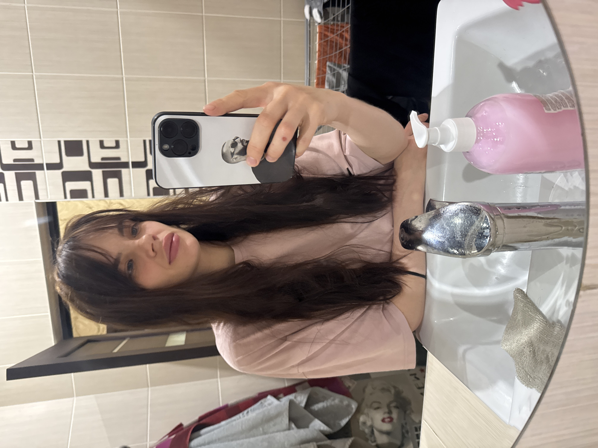 An upright mirror selfie captures a young woman in a bathroom setting. The woman, with long dark hair and bangs, is wearing a light pink t-shirt. She is holding a white iPhone, possibly a Pro model given the triple camera lenses, encased in a white and black phone case. The case features a black and white image of a bald man wearing sunglasses, resembling Walter White. Her reflection shows her looking towards the camera with light-colored eyes and pink lips.
The background consists of light beige rectangular wall tiles. Below these, a strip of decorative tiles with a repeating black and white geometric pattern is visible. To the right, a white bathroom sink holds a pink liquid soap dispenser with a white pump and a chrome faucet. A small, light-colored cloth or towel is also seen in the sink. Below the sink's reflection, an inverted image of a framed picture or poster of Marilyn Monroe with red lips is visible, along with some grey and reddish fabric. The overall scene suggests a casual moment, likely taken indoors during the day or evening with artificial lighting. No clear, legible text is visible in the image.