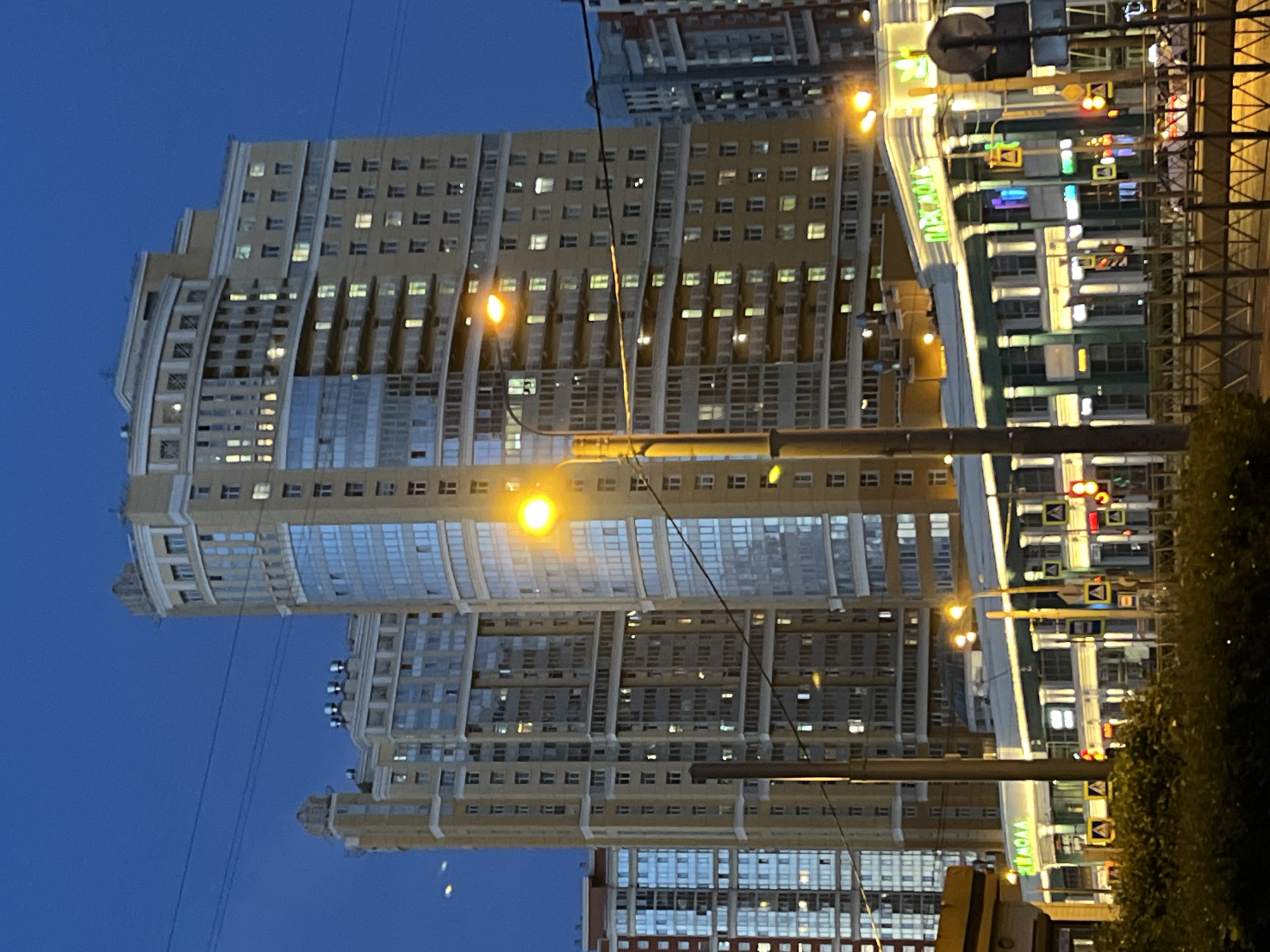 The image depicts a nighttime scene of a city street with tall buildings and streetlights. The buildings are illuminated with many windows showing interior lights, suggesting people are inside their homes. One building has a large, green sign with Cyrillic text that reads "ПЯТЁРОЧКА," which translates to "Pyaterochka," a Russian supermarket chain. Streetlights and traffic signals are visible, indicating an urban environment. The sky is dark blue, suggesting it's either dusk or nighttime. The overall impression is of a quiet, residential urban area at night.