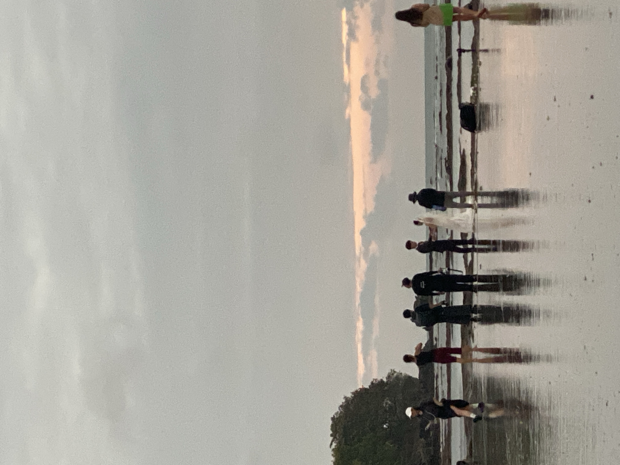 The image depicts a group of people gathered on a wet, reflective shore, likely a beach, with the ocean visible in the background. The sky is overcast with some hints of pink and orange clouds, suggesting either sunrise or sunset. On the left, a person in a white cap and dark clothing walks towards the viewer. Next to them, a person in a dark top and red shorts stands facing away. Further to the right, three figures in dark clothing stand in a line. In the center, a person in a white wedding dress with a veil is positioned next to a person in a hat and dark jacket. To the far right, a person in a light-colored top and green shorts stands facing away from the viewer. The ground is covered in shallow water, reflecting the sky and the figures present. A dark bag and a tripod are visible on the ground near the wedding couple. A dense, green treeline is present on the far left of the image. The overall scene suggests a casual gathering or possibly a wedding photoshoot on a cloudy day. The location is specified as Kuta Selatan, Indonesia.