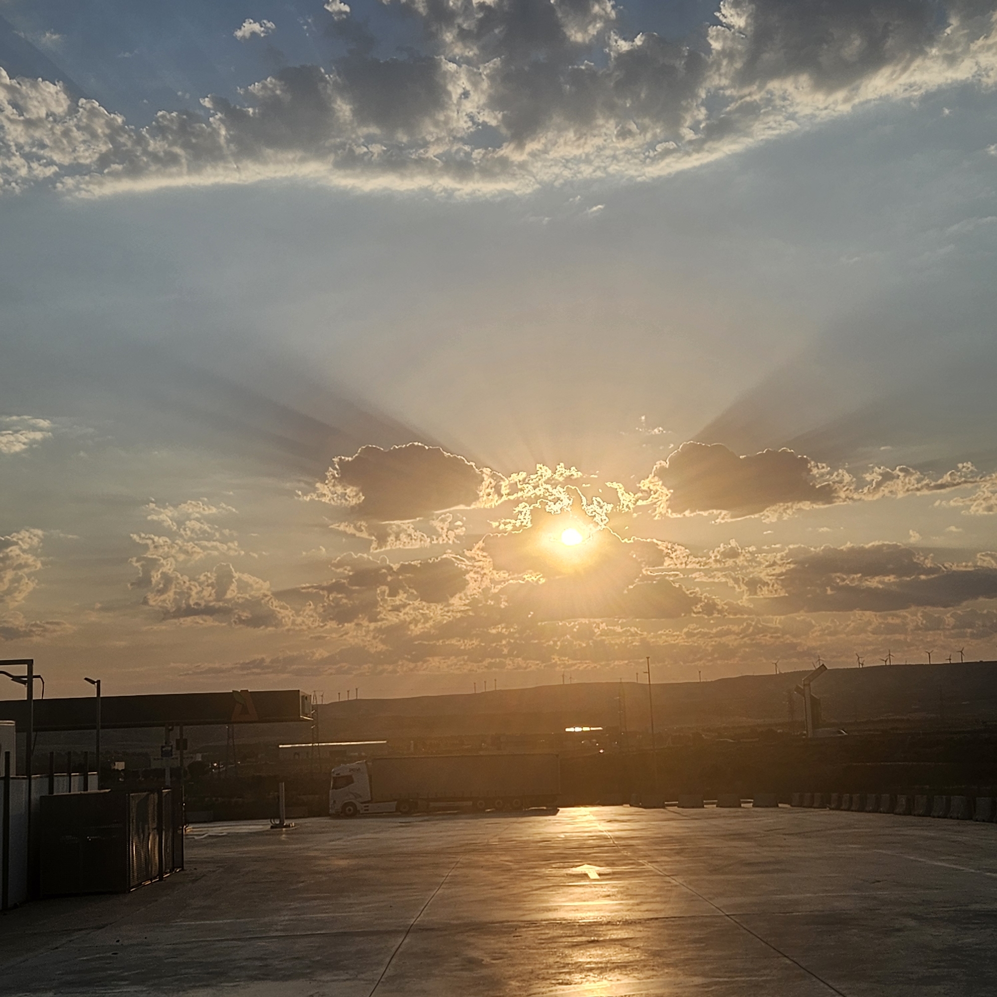 The image shows a sunset scene with the sun peeking through clouds, casting rays of light. The foreground features a paved area, likely a parking lot or industrial yard, with some storage containers on the left. A white semi-truck is parked in the mid-ground. In the background, there is a low hill with several wind turbines visible on the horizon. The sky is a mix of blue and orange, with scattered clouds. The location is identified as being in the city of Épila, Spain.