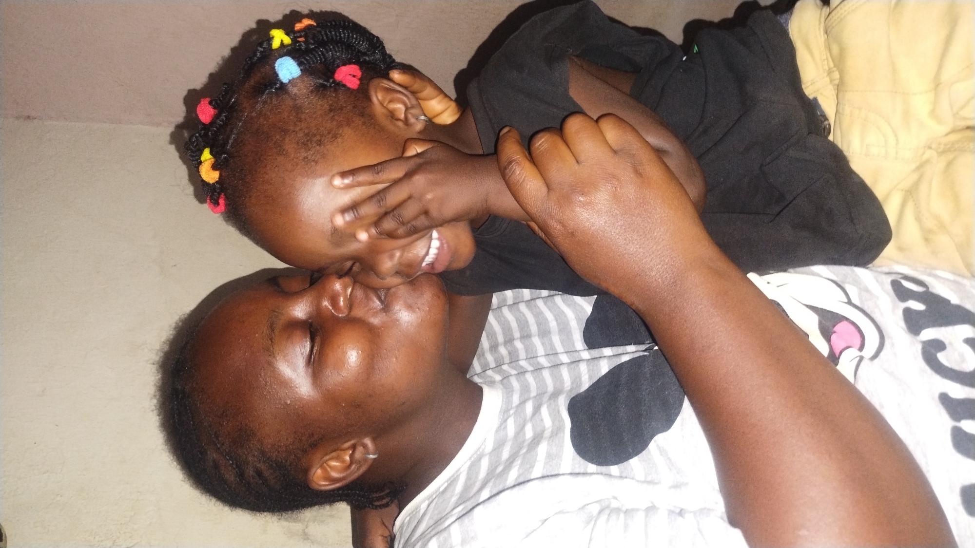 This indoor vertical photograph depicts an adult female embracing and kissing a young child, specified as being located in Jos, Nigeria. Both subjects have dark complexions and are positioned against a plain, light beige textured wall.
The adult female, positioned on the left side of the frame, has her eyes closed and lips pursed, making contact with the child's right cheek. She wears a white short-sleeved t-shirt with horizontal gray stripes, featuring a large black graphic on the chest area that includes a white shape with a pink interior, resembling a mouth. A small silver hoop earring is visible in her left earlobe. Her arms are wrapped around the child, indicating an embrace, with her right hand visible on the child's back and her left arm curving around the child's front.
The young child, positioned on the right, is smiling broadly, revealing white teeth. Their right hand is raised, with fingers splayed, covering the lower portion of their face, possibly in a playful or shy gesture. Their dark hair is braided into numerous small sections, each secured with colorful elastic bands or beads in shades of red, yellow, blue, and orange. The child wears a black short-sleeved t-shirt and light brown or khaki trousers. A small silver hoop earring is present in their right earlobe. The overall scene captures an intimate and affectionate interaction between the two individuals.