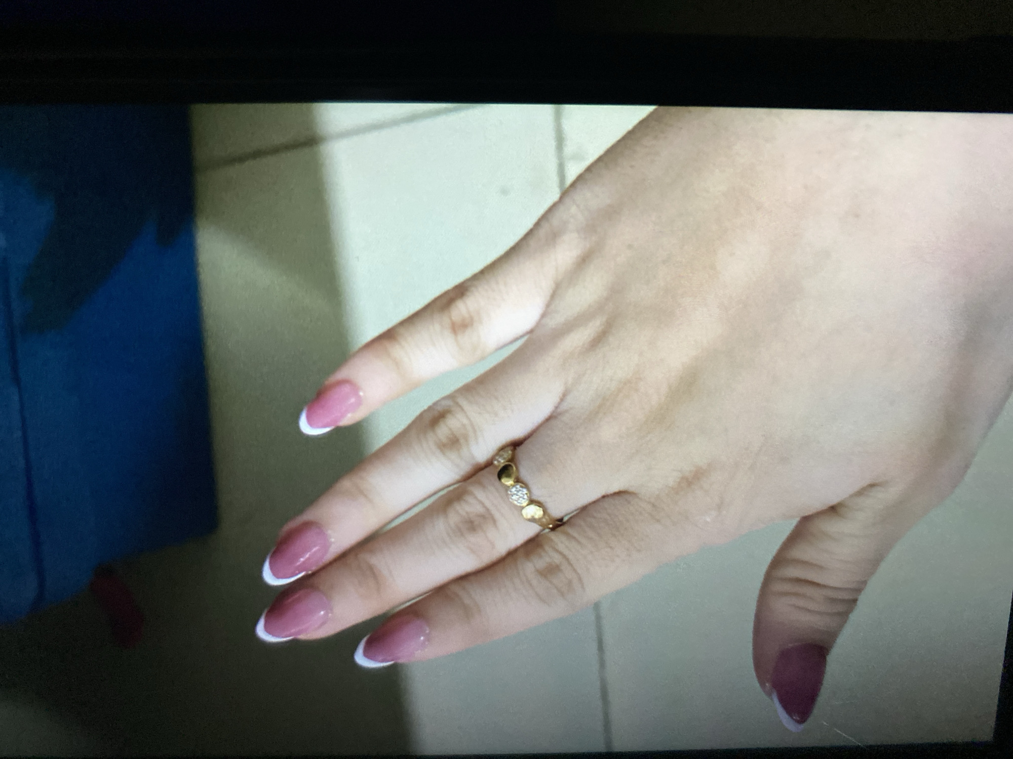 An individual's left hand, adorned with a French manicure and a gold ring featuring heart-shaped embellishments and small, clear stones, is presented against a floor and a blue object. The scene appears to be indoors, possibly in a home, given the tiling and the blue box. There are no visible elements suggesting a specific location within Ta', Indonesia, beyond it being an indoor setting.