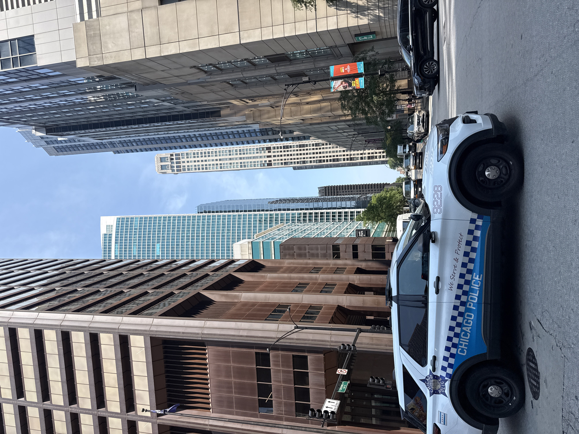 A Chicago Police SUV, vehicle number 9228, is parked on a city street. The police vehicle has "CHICAGO POLICE" written on its side, along with the motto "We Serve & Protect." Behind the SUV, tall buildings form a canyon, characteristic of a dense urban environment. These buildings vary in architectural style and materials, some featuring glass facades and others appearing older with brick or stone exteriors. Traffic signals and street signs are visible, indicating an active intersection. Other vehicles, including a black SUV and several vans, are also present on the road. The sky above is clear and blue. The scene appears to be a daytime shot taken from a slightly low angle, focusing on the police vehicle and the surrounding cityscape.