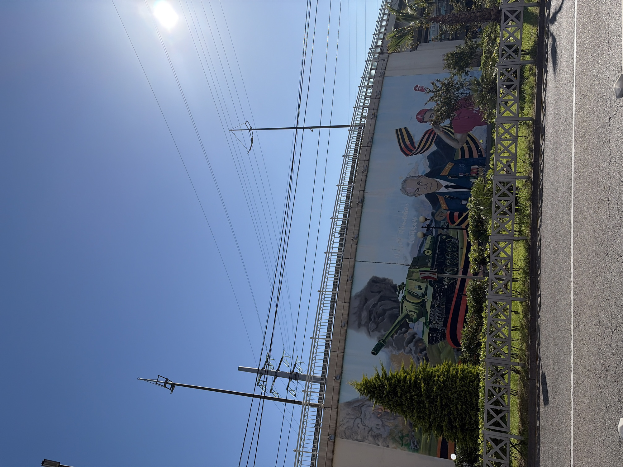 The image, taken on a clear, sunny day in Sochi, Russia, shows a large patriotic mural painted on a concrete retaining wall or bridge abutment. The mural depicts an older man with grey hair in a dark blue military uniform adorned with medals, resembling a high-ranking officer or general. To his right, a younger figure wearing a red cap and shirt holds a prominent black and orange striped St. George ribbon, a symbol of military valor. Below these figures, a green tank is painted amidst smoke and debris. Visible Cyrillic text on the mural includes "Победы" (Victory) and "75 лет" (75 years), suggesting a commemoration, likely of the 75th anniversary of Victory Day (World War II).
In front of the mural, a strip of vibrant green grass and shrubs is bordered by a decorative grey metal fence. To the right of the fence is an asphalt road with a distinct white lane marking. Overhead, numerous power lines crisscross the bright blue sky, supported by tall utility poles. The sun is high, casting a strong flare in the upper left of the frame. Palm trees are visible growing on top of the wall structure, adding to the southern climate aesthetic. The scene captures an urban roadside environment, with the mural serving as a prominent public art piece celebrating military history.