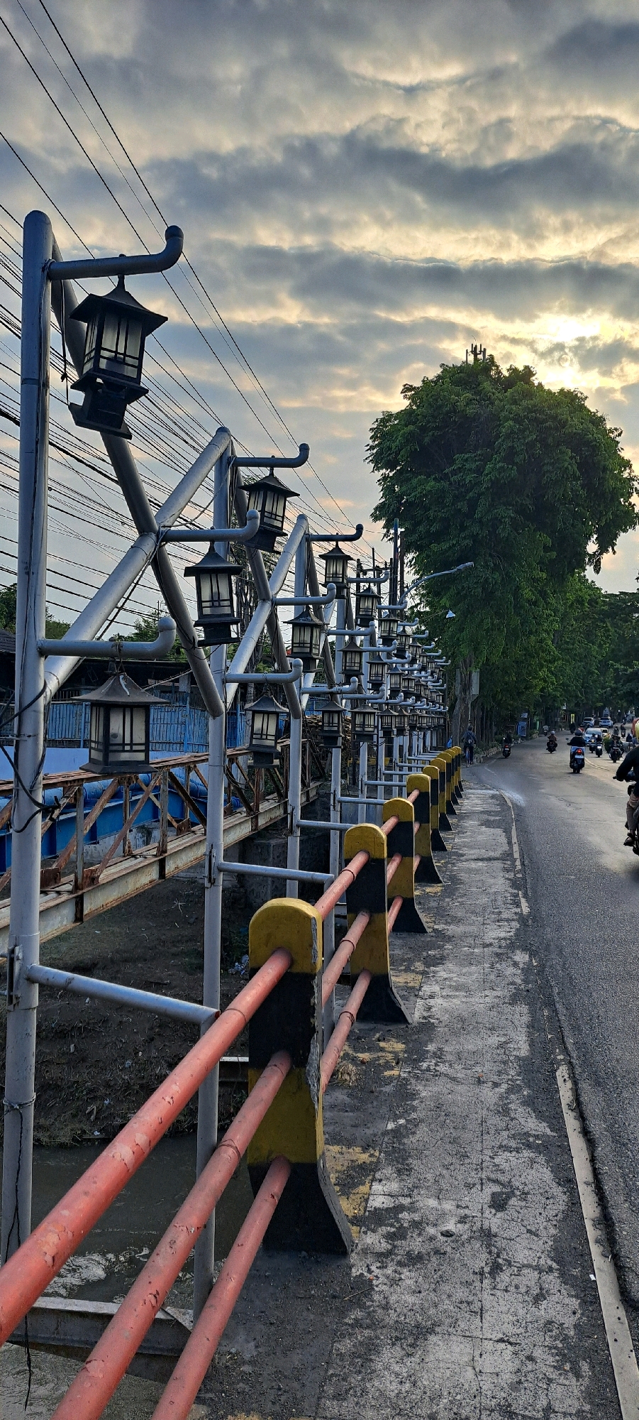The image depicts a street scene in Pasuruan, Indonesia, captured during the day with cloudy skies. The foreground features a sidewalk with a railing, characterized by red and yellow horizontal bars and black supporting pillars. Beyond the railing, a bridge with a blue structure is visible, showing signs of rust on its metal components. The sidewalk is adjacent to a road where several motorcycles are being ridden. The left side of the image is lined with a series of decorative streetlights, mounted on a metal structure with multiple arms, each holding a lantern. Overhead, a dense network of electrical wires crisscrosses the sky. In the background, a large, leafy green tree dominates the right side of the frame, casting shadows. Further down the road, more vehicles, including cars, are visible. The overall impression is an urban environment with a blend of infrastructure, transportation, and natural elements.