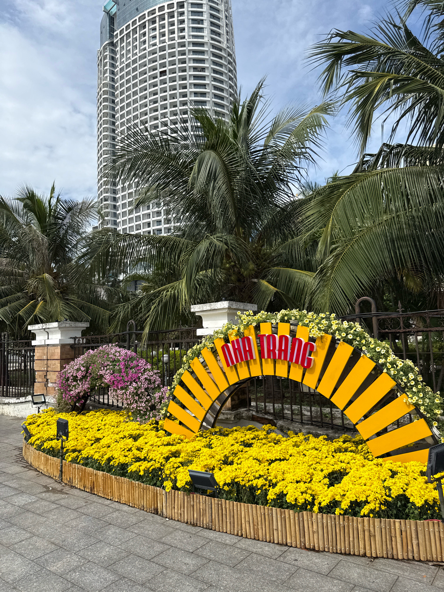 A tall, white, modern skyscraper with many windows dominates the background of the image. In front of the skyscraper, lush green palm trees are visible. A semicircular, yellow archway decorated with small white flowers and red lettering that spells "NHA TRANG" is a prominent feature in the foreground. This archway is situated in front of a black metal fence, behind which are bushes with pink flowers. The archway is surrounded by a bed of vibrant yellow flowers, bordered by a bamboo fence. The ground in front of the flowers is paved with gray square tiles. Several black spotlights are placed around the flower bed. The sky is cloudy and blue.