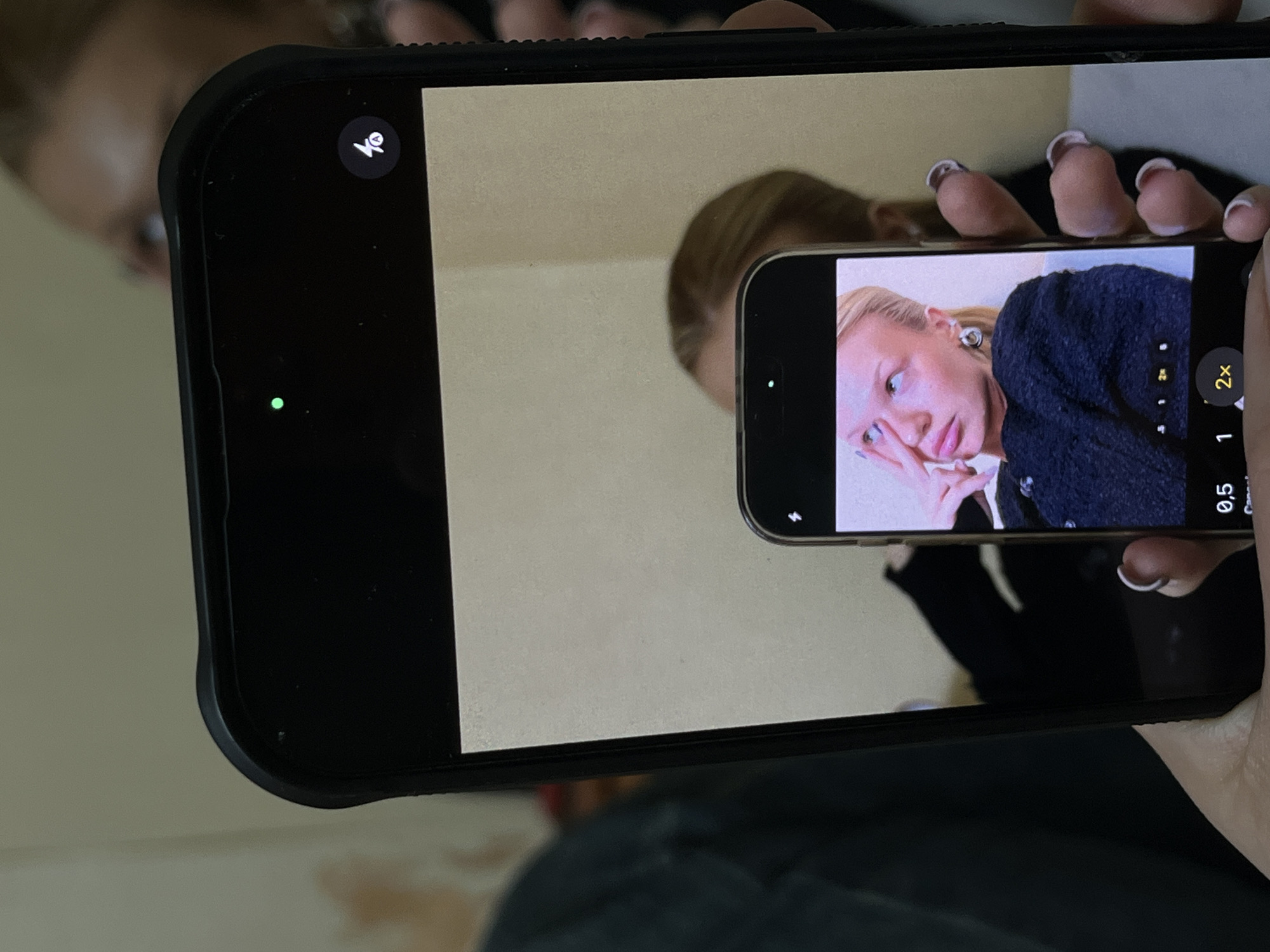 A person is holding a black iPhone to take a picture of another iPhone, which is displaying a woman posing with her hand to her face. The scene appears to be indoors. The location in the city of Chaikovsky, Russia, is not identifiable from this image.