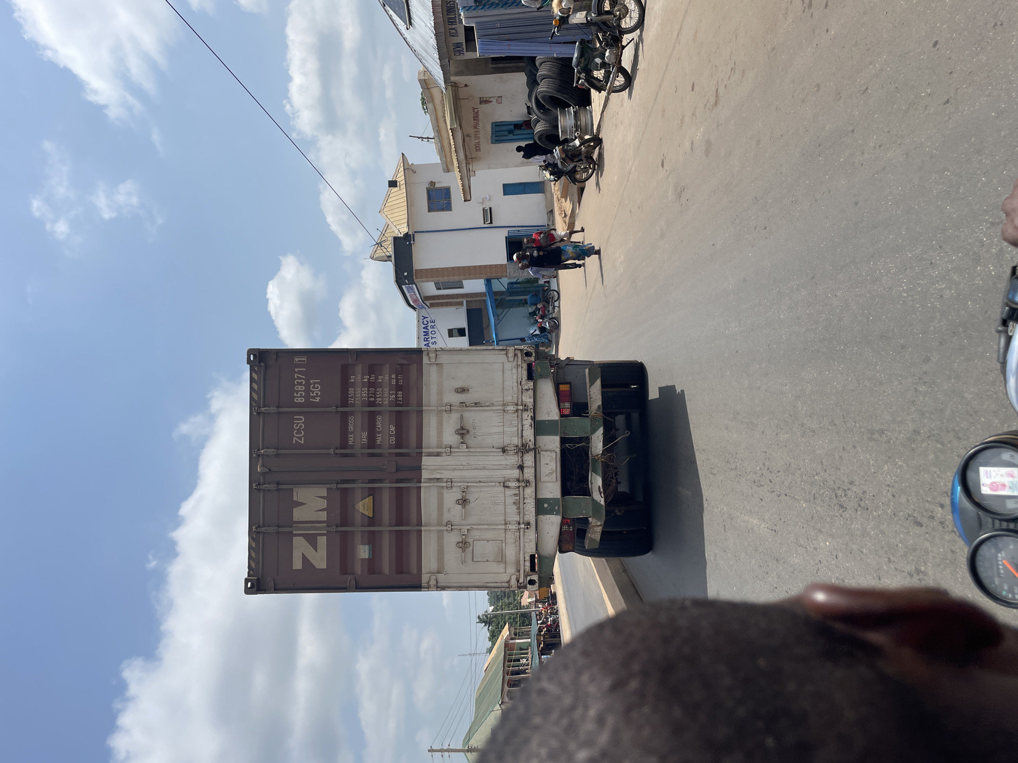 The image depicts a scene on a street in Akwanga, Nigeria, under a partly cloudy sky. In the foreground, the rear of a person's head is visible, suggesting the photo is taken from a moving motorcycle. The motorcycle's dashboard with a speedometer is also partially visible.
Dominating the mid-ground is a large, maroon shipping container on a truck. The container displays the logo "ZIM" and identification numbers "ZCSU 858371 1 45G1", along with weight and volume specifications.
To the right of the truck, a street scene unfolds. Several buildings line the street, including one with a sign that reads "PHARMACY STORE" and another with "ROYAL HOPE PHARMACY". Large stacks of tires are visible outside one of the buildings. People are walking on the street, and several motorcycles are parked or in motion.
The road surface is asphalt, with some markings and debris. The architecture of the buildings suggests a local style, with some appearing to be shops or commercial establishments. The overall impression is a busy street in a Nigerian town.