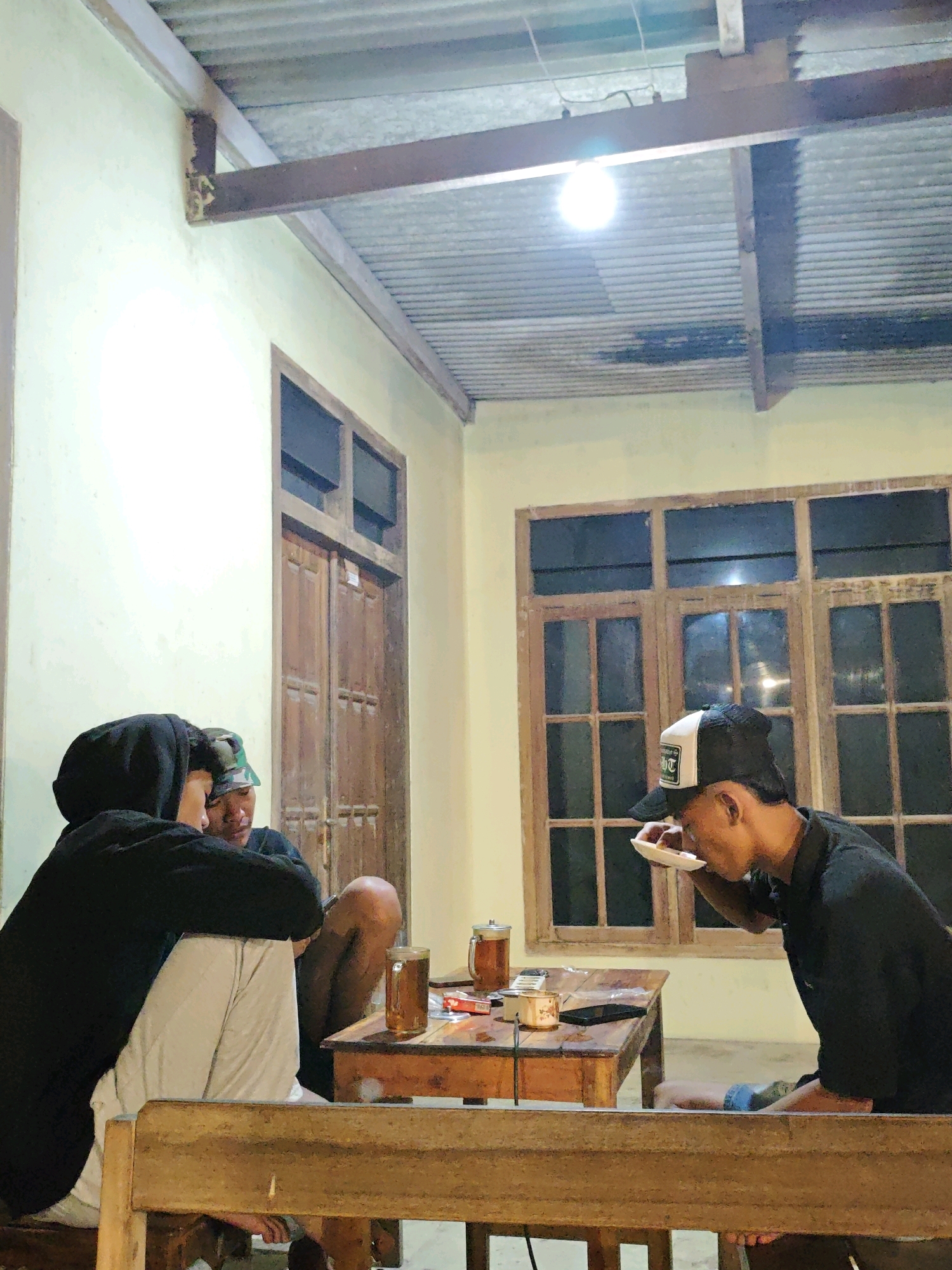 In Ngasem, Indonesia, three young males are seated around a wooden table under a corrugated metal roof supported by exposed wooden beams. The room appears to be an open-air structure or a covered patio. Two individuals in the foreground are positioned to the left, facing the table. One, wearing a black hoodie and light-colored pants, is seated and leaning forward. The other, with a camouflage hat, is seated next to him, also looking towards the table. A third individual, on the right, wears a black t-shirt and a black baseball cap with a white logo. He is actively eating from a white plate. On the table are two glasses of amber liquid, possibly tea, a cell phone, and an open pack of cigarettes. The background features a wooden double door and a large window with multiple panes, suggesting a residential or semi-outdoor commercial establishment. The lighting is dim, with a single bright bulb overhead.