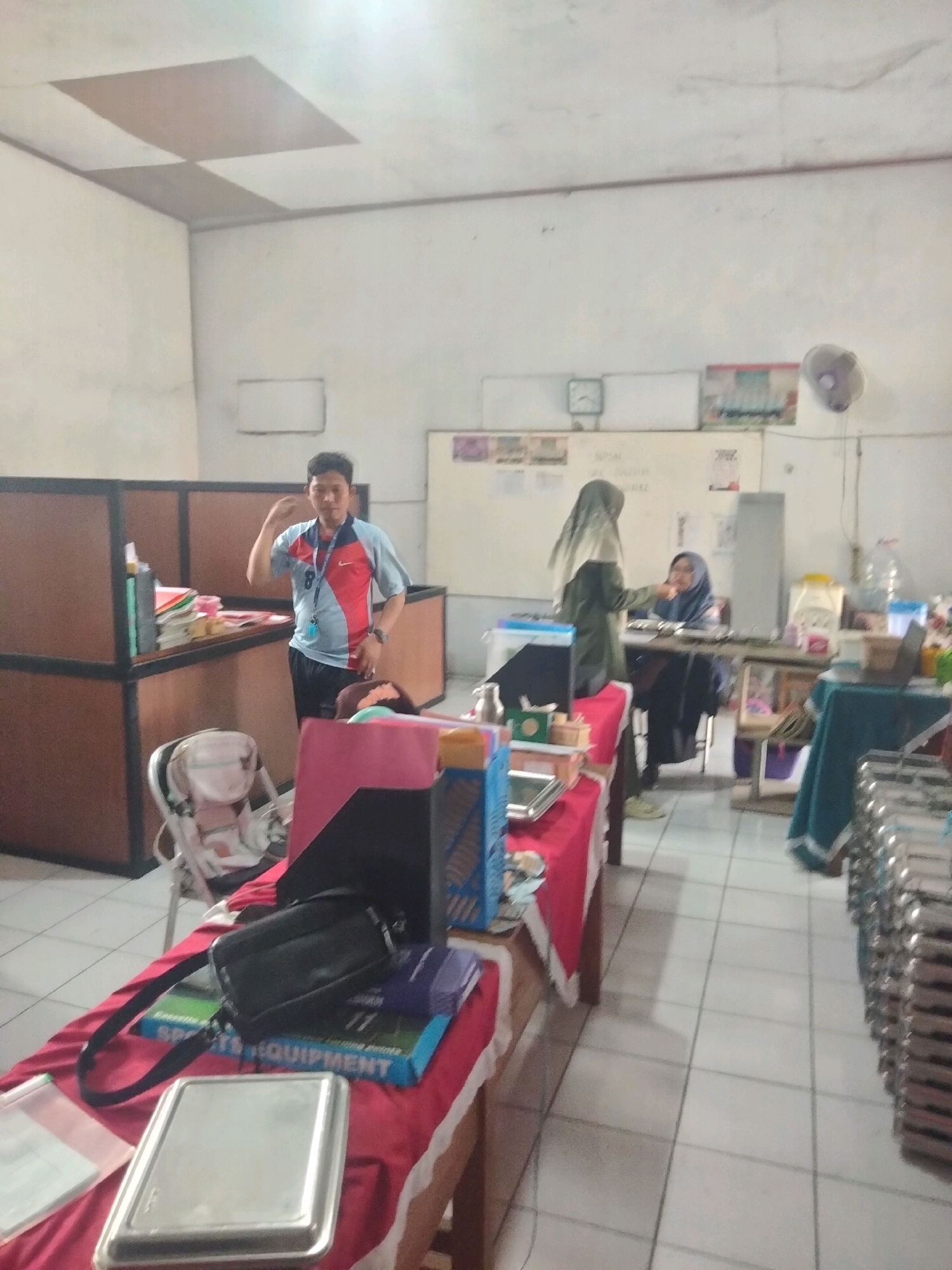 In Suka Sari, Indonesia, a dimly lit indoor room appears to be a classroom or office space. The room has white walls and a tiled floor. A man in a red and blue shirt stands near a partition. In the background, two women are seated at a desk. One woman, wearing a hijab, is interacting with the other. Various objects are scattered around the room, including tables, chairs, papers, and books. The overall scene suggests a casual, possibly academic setting.