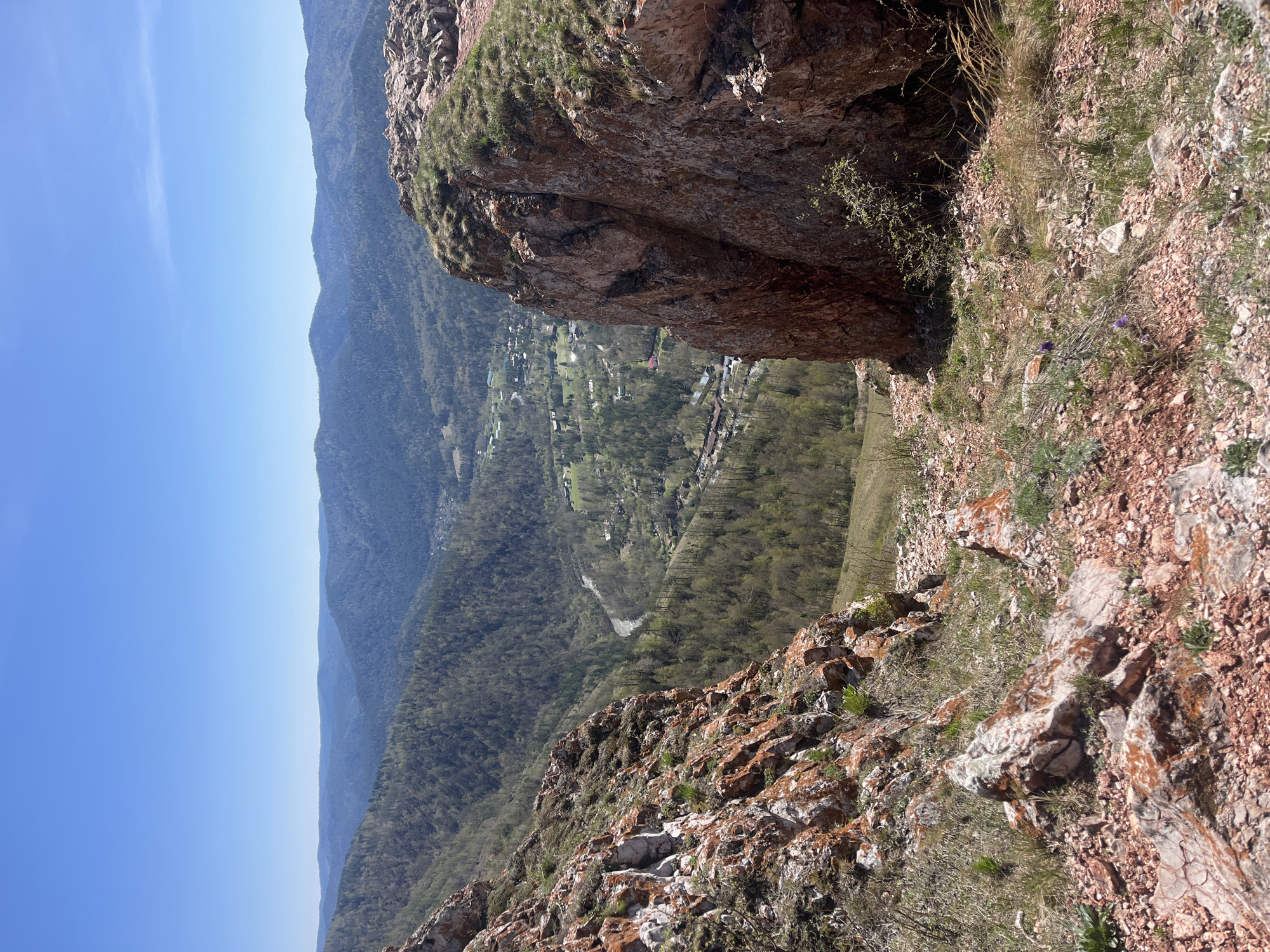 The image depicts a mountainous landscape, likely taken from a high vantage point. In the foreground, rocky terrain with sparse vegetation is visible. A large rock formation dominates the right side of the frame. Below, a valley unfolds with trees and a small settlement visible in the distance. Beyond the valley, a series of mountain ranges fades into the horizon. The sky is clear blue. Based on the location of Krasnoyarsk, Russia, the image likely shows the Stolby Nature Reserve area.
