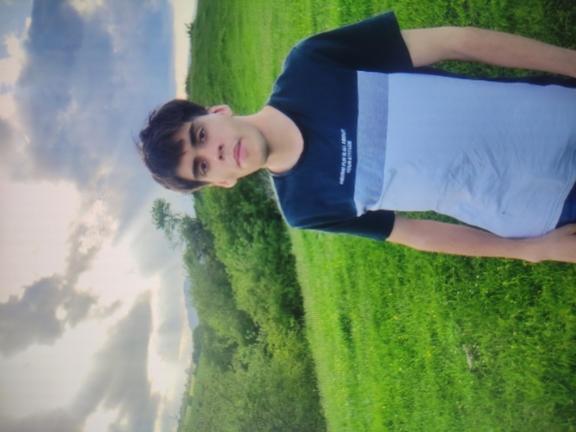 A young man with dark hair and a light complexion stands in a grassy, hilly area. He is wearing a t-shirt with dark blue sleeves and collar, and a light blue and white striped body. The background consists of rolling green hills covered in grass and trees, with a cloudy sky overhead. There is no indication of the city of Baku, Azerbaijan in the image.
