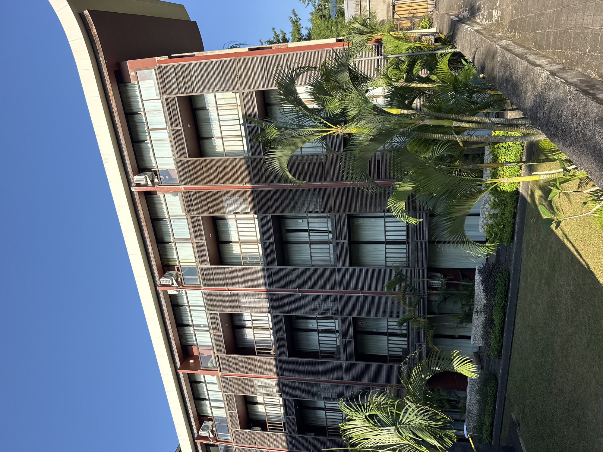 The image shows a multi-story building in Kuta Selatan, Indonesia. The building has a modern architectural style with horizontal wooden slats covering the exterior walls. The windows are visible behind the slats, with white curtains. There are small air conditioning units installed outside some of the windows. Palm trees are planted in front of the building, and a grassy lawn extends to the side. A concrete wall or walkway is visible at the edge of the lawn. The sky is blue, and the sunlight indicates it is daytime.