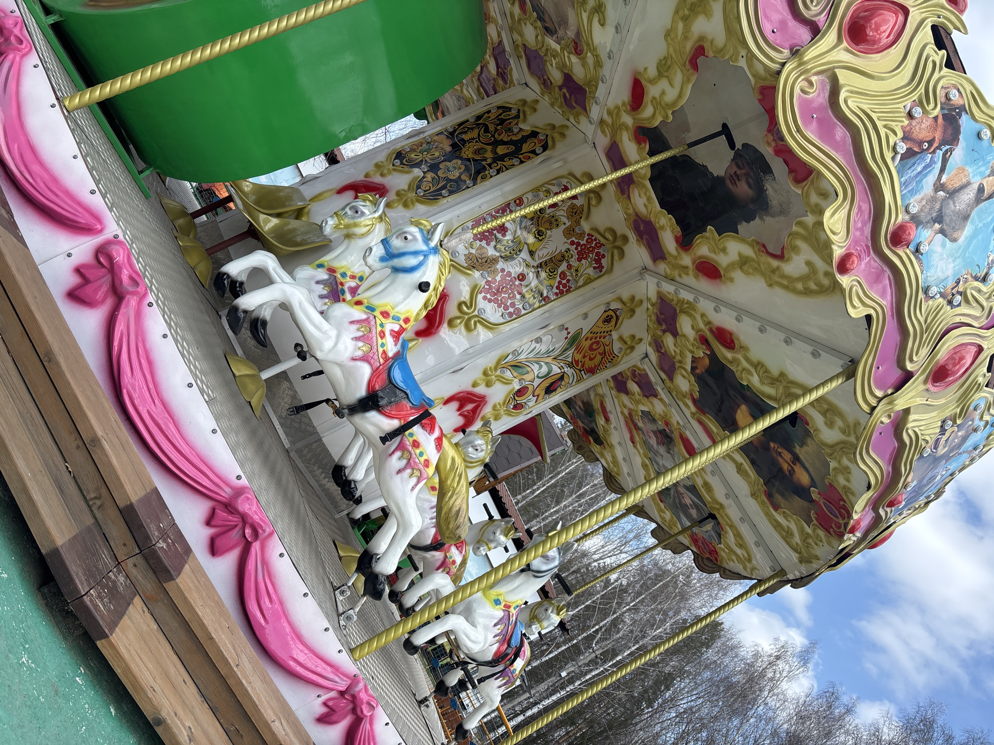 The image shows a carousel in the city of Chaikovsky, Russia. The carousel has white horses with colorful saddles and bridles. The horses are arranged around a central platform with a green canopy. The carousel's decorative panels feature paintings of people and animals, as well as floral patterns. The structure is decorated with pink and gold accents. In the background, bare trees and a blue sky with clouds are visible. The carousel is located outdoors.