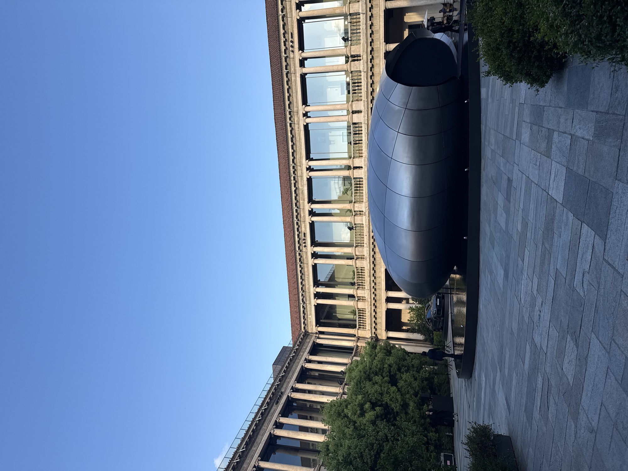 An outdoor urban scene in Milan, Italy, features a grand building, a sleek modern sculpture, and a clear blue sky. The image, originally rotated, depicts a bright day.
On the left, a vast expanse of clear, bright blue sky dominates, indicating fair weather. To the right and extending across the middle, a large, multi-story building with a classical or neoclassical architectural style is prominent. It features numerous columns, balustrades, and large windows that reflect the sky and faint clouds. The building's facade is made of light-colored stone, and a reddish-brown roof line is visible at its top. Below the building, green trees are visible, adding natural elements to the urban landscape.
In the foreground, a striking dark, metallic sculpture sits on a paved plaza. The sculpture is elongated and smooth, resembling a large, futuristic capsule or an abstract, segmented pod. It appears to be resting in a dark, shallow water feature or on a reflective base, which shows faint reflections of the sky and a dark vehicle. The ground in the foreground is paved with light grey rectangular stones arranged in a grid pattern. A neatly trimmed green bush is located behind the sculpture on the right.
A few indistinct figures can be seen walking on the paved area and in the distance, suggesting a public space. The strong, clear daylight indicates it is daytime, likely morning or late afternoon. No discernible text is visible in the image.