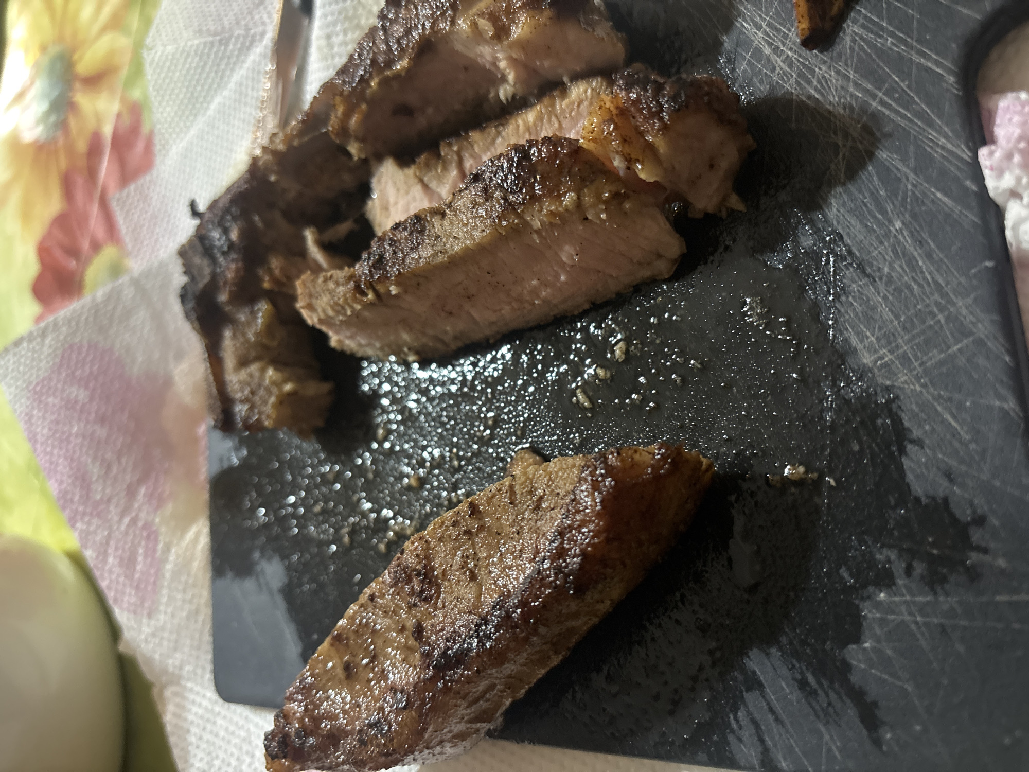 The image shows slices of cooked steak on a black cutting board. The steak appears to be medium-rare with a browned exterior. Juices from the steak are visible on the board. The background includes a floral-patterned napkin and a white object. There is no information in the image to determine a location in Goritsa, Bulgaria.