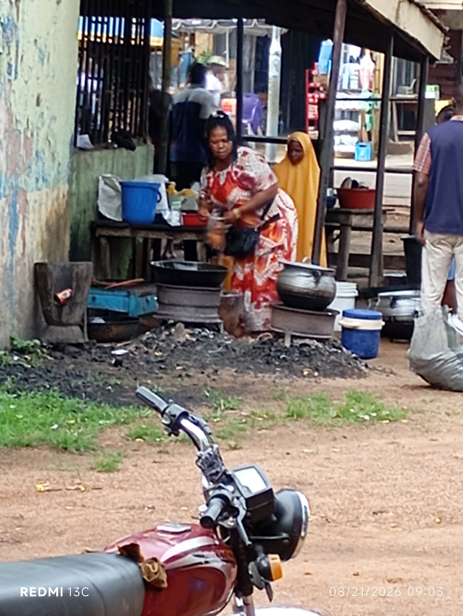 A street food stall is set up outdoors in Shendam, Nigeria. Two women are visible, one in the foreground wearing a red and white patterned dress, and another in the background wearing a yellow hijab. Behind them, various cooking pots and equipment are arranged on a table and on the ground, including what appears to be a mortar and pestle. The stall is situated near a building with weathered walls and a metal grate. In the foreground, the front of a red motorcycle is visible. The ground is a mix of dirt and grass, with scattered debris. The time stamp "08/21/2026 09:03" is displayed at the bottom right of the image.