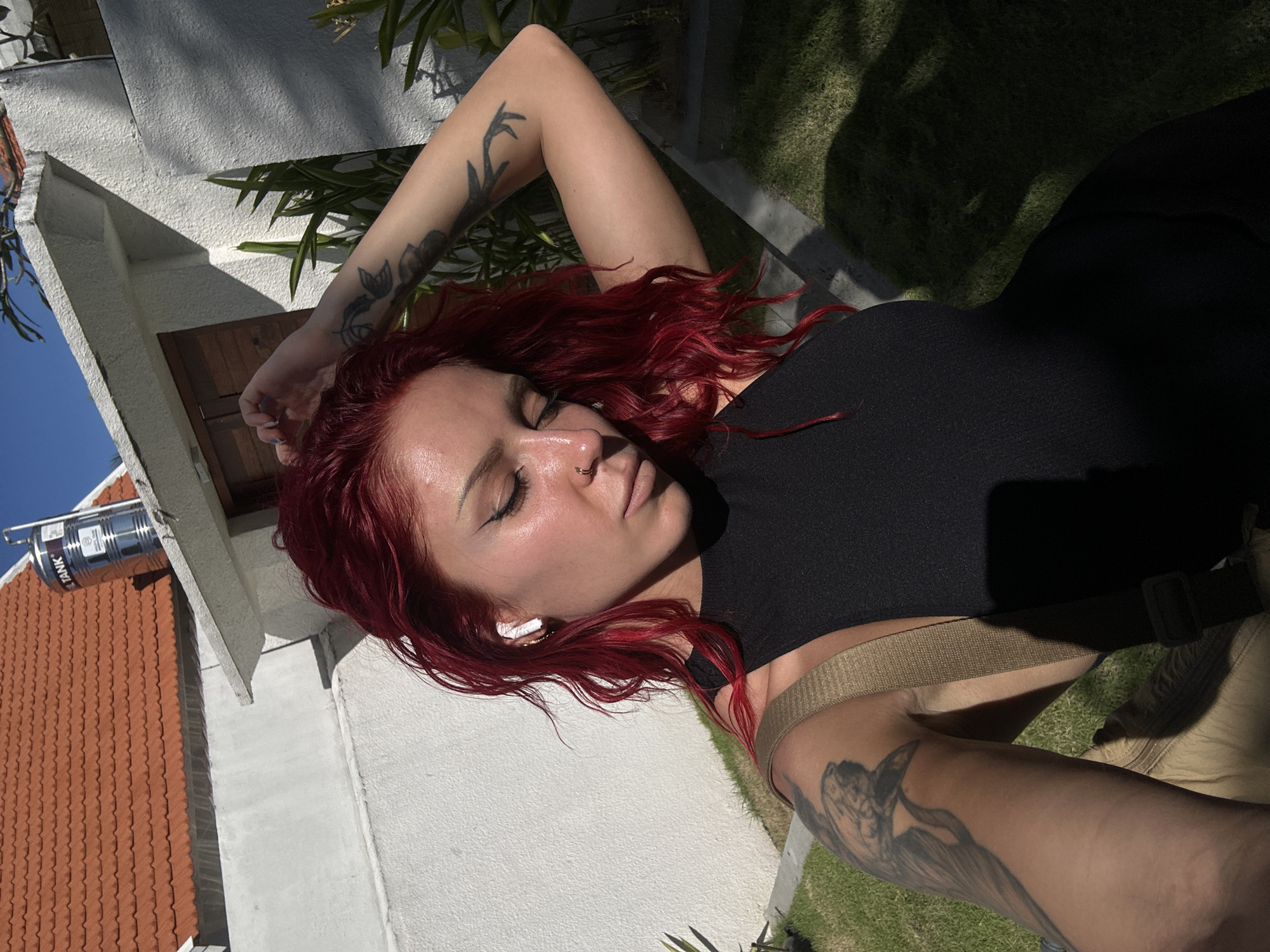 The image shows a woman lying on a surface with grass and a building visible in the background. The woman has red hair, closed eyes, a nose ring, and an earbud in her left ear. She is wearing a black top and a light-colored belt. She has tattoos on both arms. The building has a white wall, a red tile roof, and a metal container attached to it. The location is in North Kuta, Indonesia.
