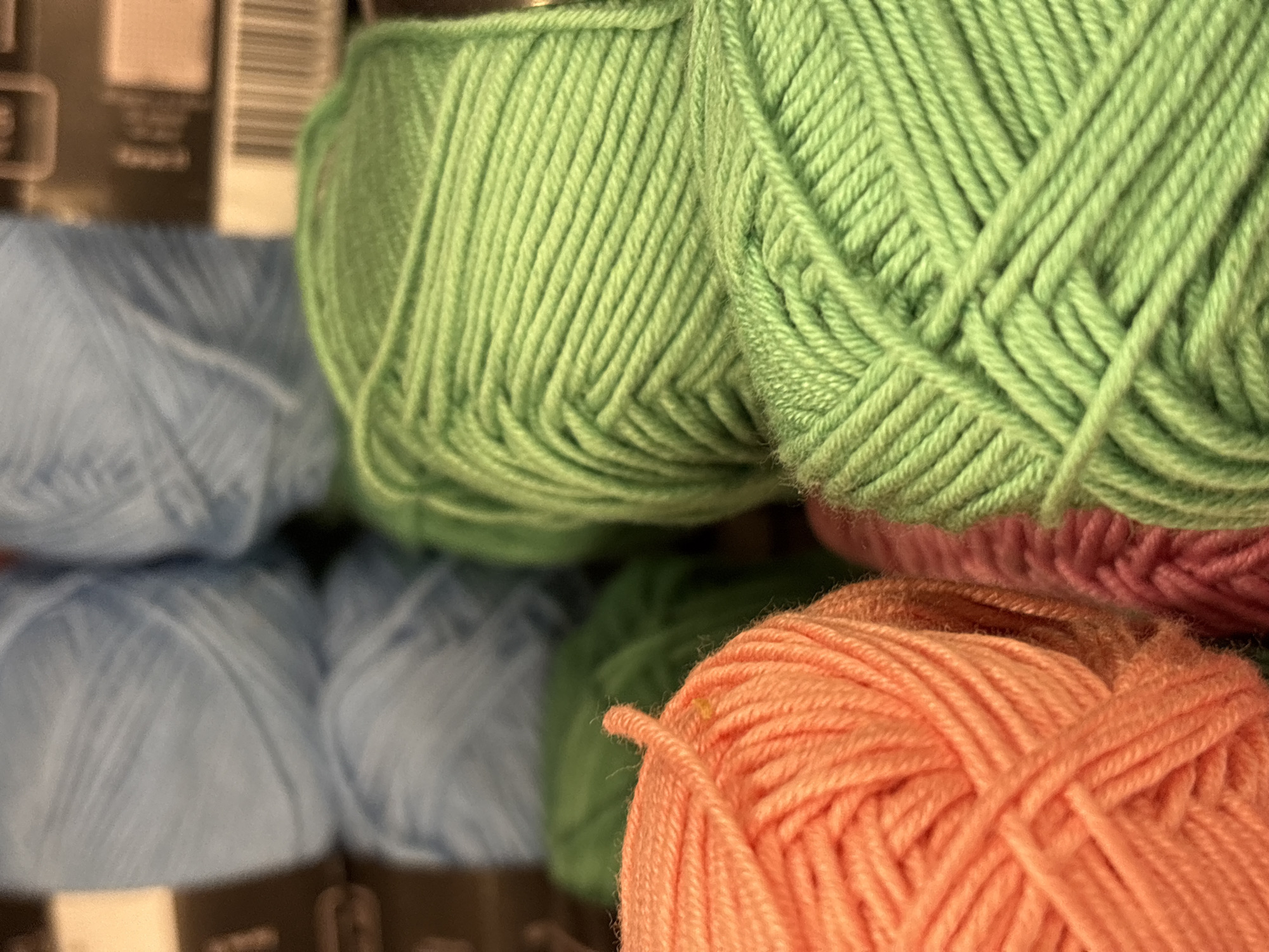 This media file features a close-up, eye-level shot of several balls of yarn stacked in a retail setting. The foreground is dominated by a vibrant ball of peach-colored yarn, with some finer threads beginning to unravel from its surface. Behind it, partially visible, is a ball of pink yarn. Above these, and further into the background, are two large balls of bright green yarn. To the left and slightly out of focus are several balls of light blue yarn, also stacked.
The image has a shallow depth of field, with the peach and green yarns being the sharpest. The texture of the yarn is highly detailed, showing the individual strands and the way they are wound into balls. The lighting appears to be artificial, consistent with an indoor retail environment. There are no people visible. The overall impression is one of colorful craft supplies.
On the far left edge of the image, partially obscured by the blue yarn, are elements of packaging that resemble the labels on yarn products. Some text is visible, appearing as horizontal lines and what might be barcodes, but it is too blurry and incomplete to be deciphered. There are no overt cues to the time of day, weather, or specific emotions, beyond the visual appeal of the bright colors. The presence of multiple stacks of yarn suggests a store that sells craft or hobby supplies. Given the location context, this is likely a shop in Søborg, Denmark.