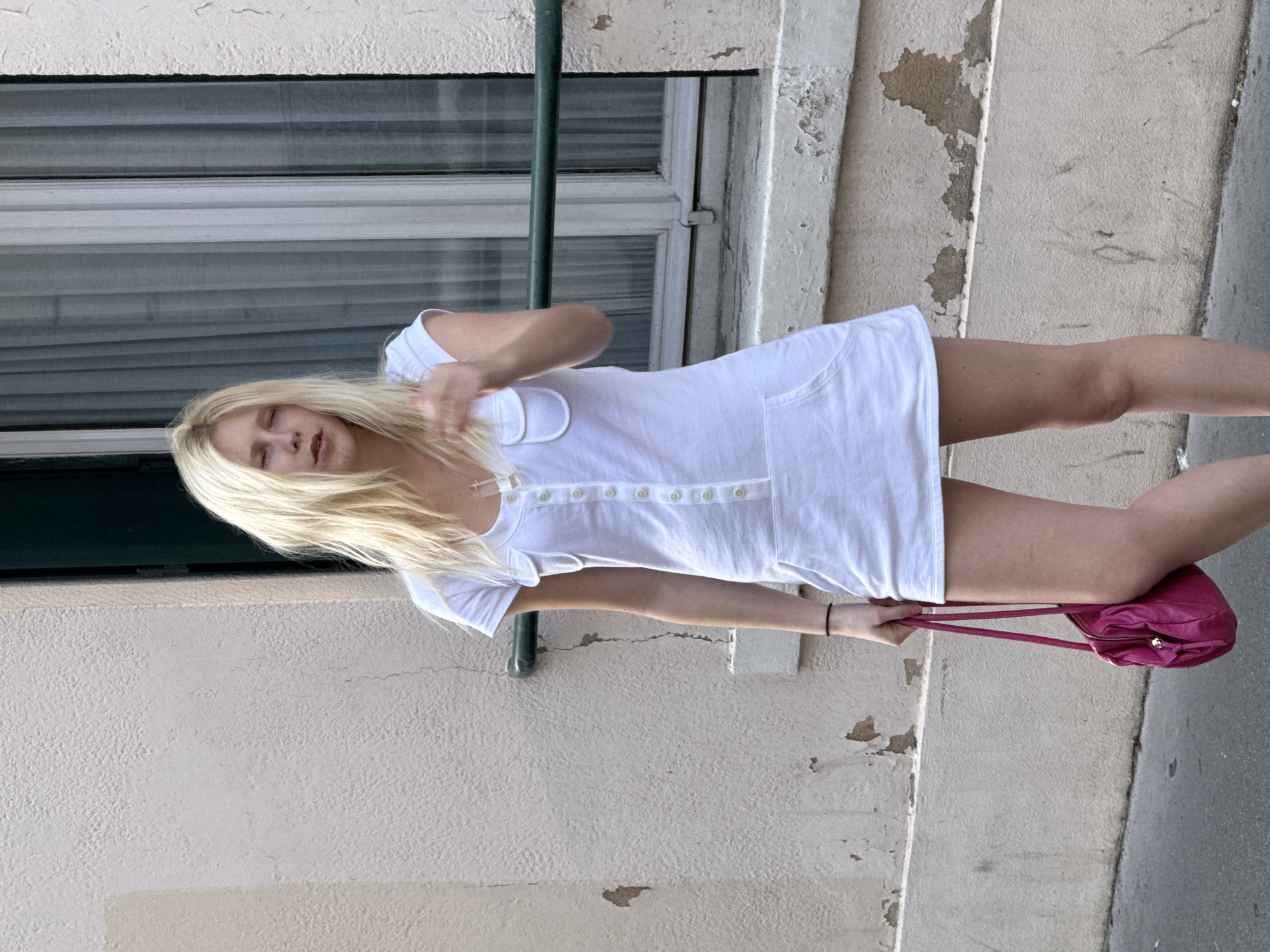 A woman with blonde hair is standing on a sidewalk in front of a building. She is wearing a short white dress and holding a small pink handbag. The building has a window with blinds. The location is likely Paris, France, given the architectural style.