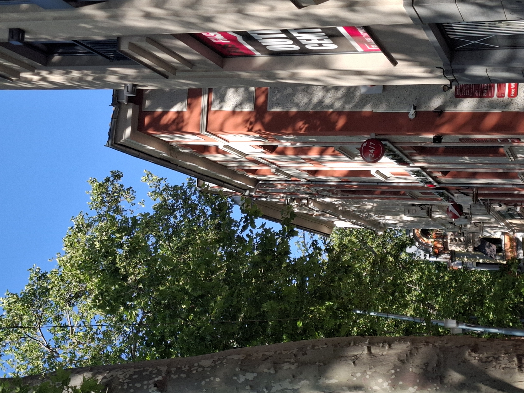 A low-angle, full outdoor shot shows a tree on the left, a building with red and white accents in the middle, and another building with a window sign on the right. The sky is clear and blue. The tree trunk on the left is light brown with a dappled texture. Lush green foliage fills the upper left and middle portions of the image. The building in the middle features red-painted sections and white decorative trim. A circular red sign that reads "24/7" is visible on its facade. The building on the right is a lighter color and has a large window with a black and red sign that reads "FASHION" and "OUTLET CENT". Shadows from tree branches are cast across the buildings. A streetlamp is present between the tree and the middle building.