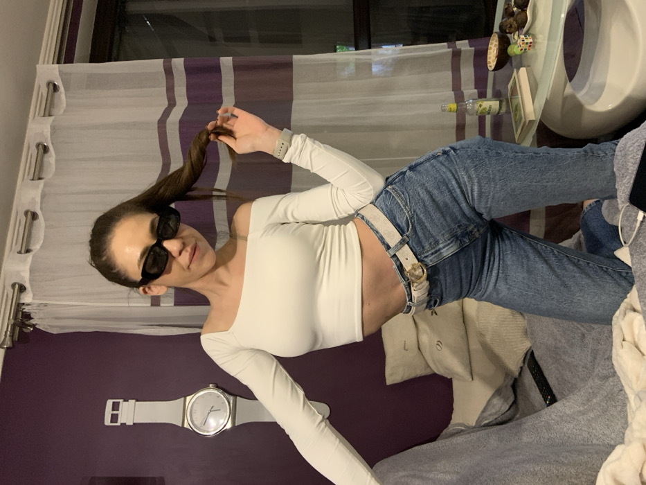A female subject is depicted indoors, kneeling on a grey textured blanket. She wears a white off-the-shoulder crop top, light blue denim jeans, a light-colored textured belt with a gold-colored buckle, and black rectangular sunglasses. Her dark hair is pulled into a high ponytail, which she holds with her left hand, revealing a light grey smartwatch on her wrist. The background features a dark purple wall to the left, adorned with a large white wristwatch-shaped wall clock indicating approximately 10:10. To the right, white sheer curtains with horizontal purple stripes obscure a window showing a dark exterior. Stacked pillows are visible behind the subject on the left, and a black television remote control rests on the blanket nearby. A low white table to the subject's right displays a small clear bottle with a yellow label and a lemon slice, a light-colored book, a coconut shell bowl containing dark spherical items (likely chocolates or truffles), and a multi-colored puzzle cube. A charging cable and part of a dark smartphone are visible on the blanket near the table. The specific location within Sofia, Bulgaria, cannot be determined from the visual information provided.