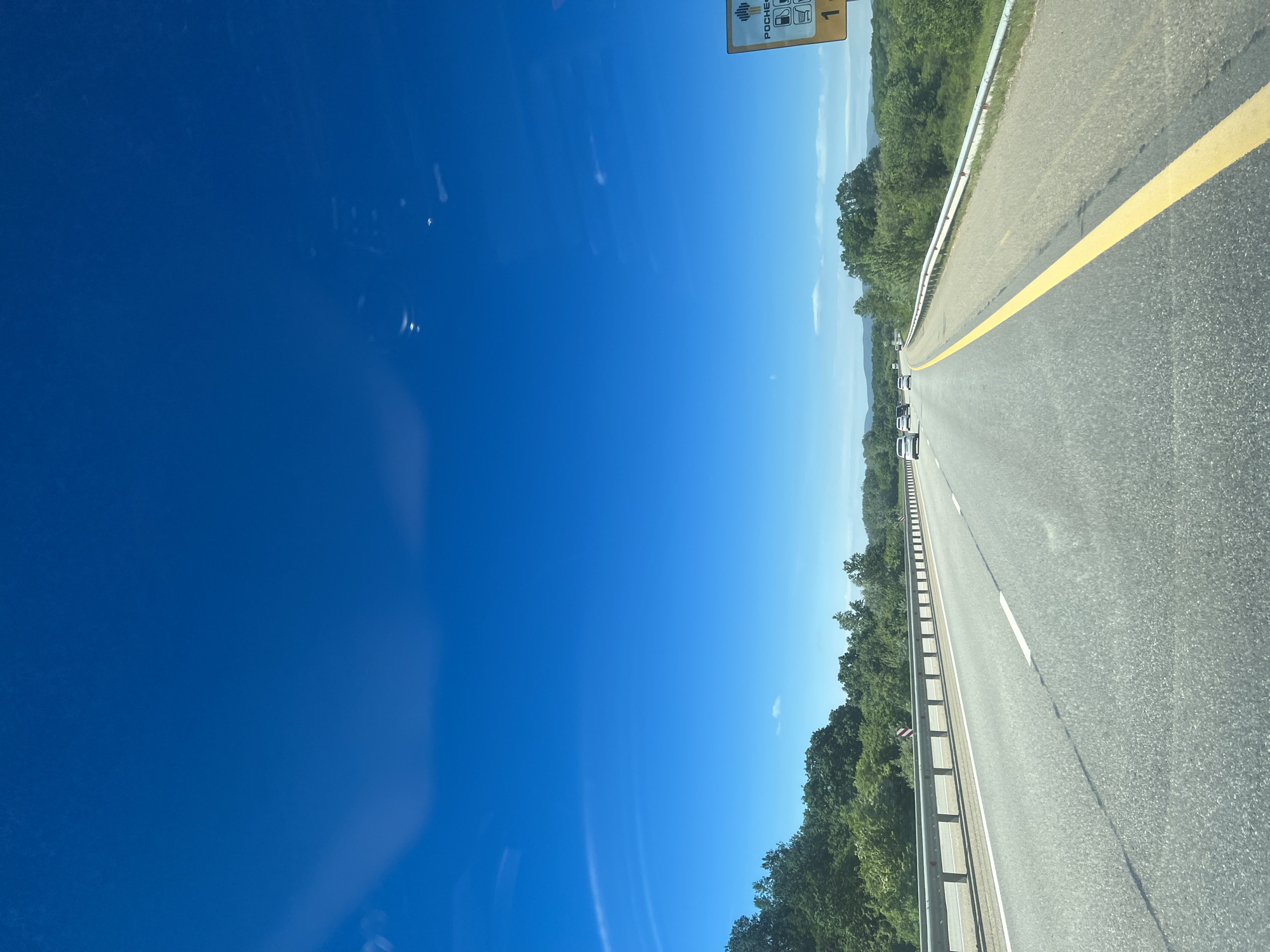 The image depicts a view from a vehicle traveling on a highway in Saratovskaya Oblast, Russia. The scene captures a straight section of the road with two lanes in each direction. A solid yellow line divides the lanes, and a metal guardrail runs along the roadside. The road is lined with trees. Several vehicles are visible in the distance on the same lane. The sky is clear and blue. A highway sign can be seen in the top left of the frame.