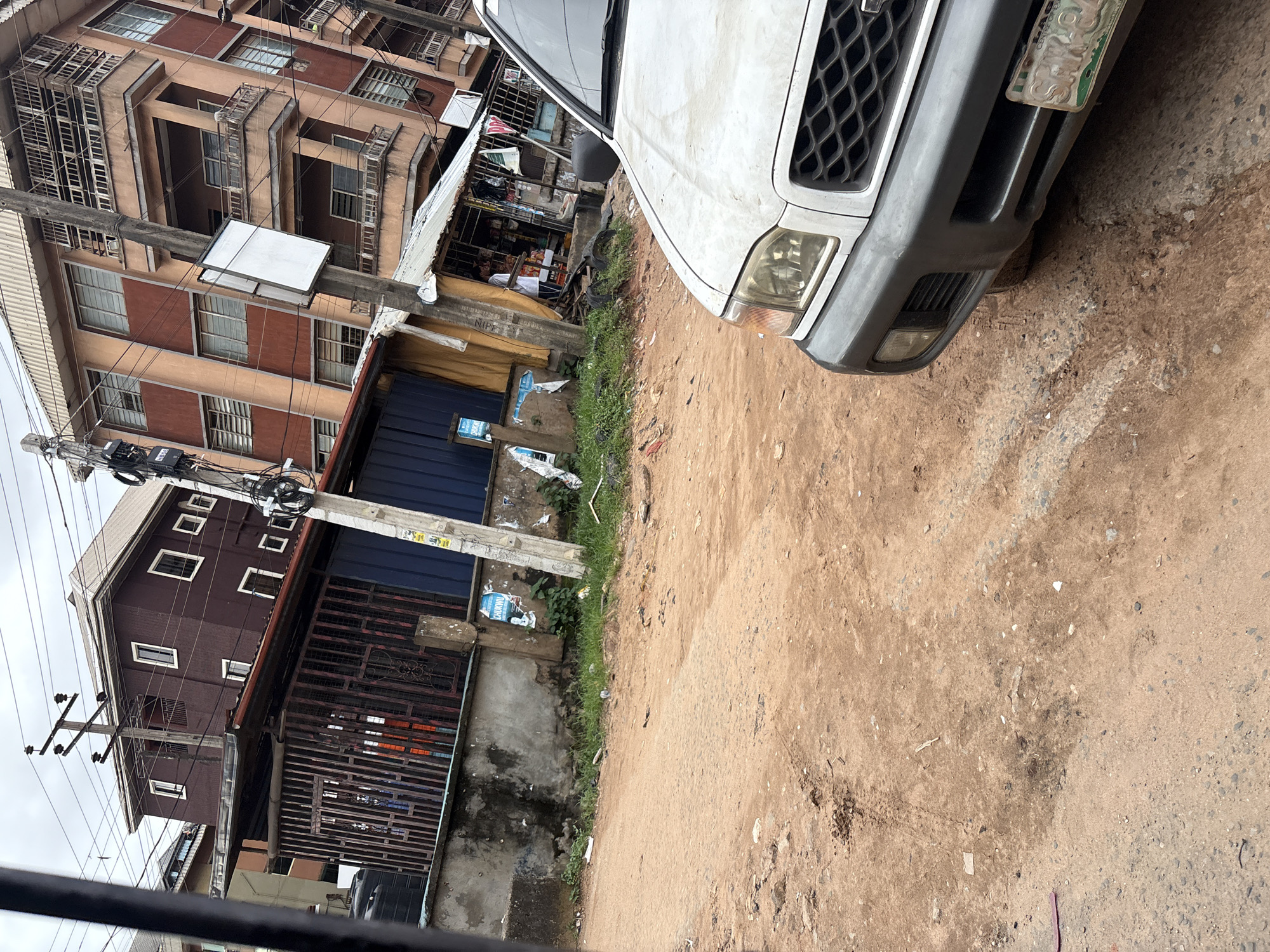 The image captures a street scene in Nkpor, Nigeria, dominated by a dirt road and several buildings. In the foreground, the front of a white vehicle is visible, showing its bumper, grille, and part of the headlight. The vehicle is positioned on the right side of the frame, facing left.
The road surface is uneven and made of reddish-brown dirt, with visible tire tracks and scattered debris. To the left of the road, a concrete wall runs along the edge of a property. Behind this wall, a dark metal gate encloses an area with some items visible inside, including what appears to be laundry hanging. Adjacent to this is a blue corrugated metal structure.
Further back, a multi-story building with a dark brown facade and white-framed windows stands prominently. It has a slanted roof and is adorned with numerous electrical wires and poles. Another taller building, with an orange and brown exterior and multiple balconies with metal railings, is located to the right of the dark brown building. A small, open-air shop with a metal roof and various goods displayed is situated in front of the orange and brown building. A person can be seen standing in the shop doorway. The overall environment suggests an urban setting with mixed-use buildings and commercial activity. The sky is overcast, indicating a cloudy day.