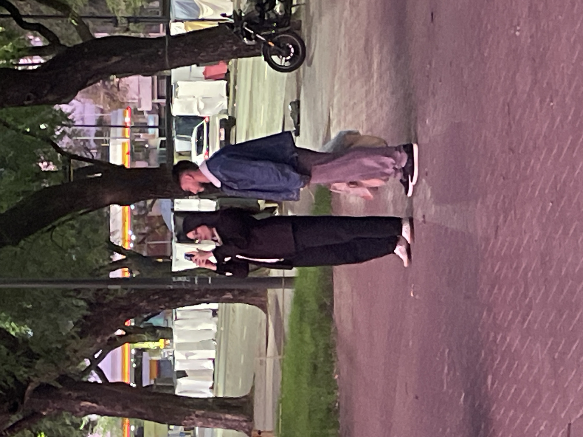 Two individuals are standing on a sidewalk in a city. One individual, wearing a dark hooded jacket, dark pants, and white shoes, is looking at a phone. The other individual, wearing a blue denim jacket, brown pants, and dark shoes with white soles, is standing next to the other. The sidewalk is next to a street with parked cars, grass, and trees. There is a motorcycle parked on the street. In the background, there are buildings and trees. Based on the architectural style and presence of street lights, this could potentially be Buenos Aires, Argentina.