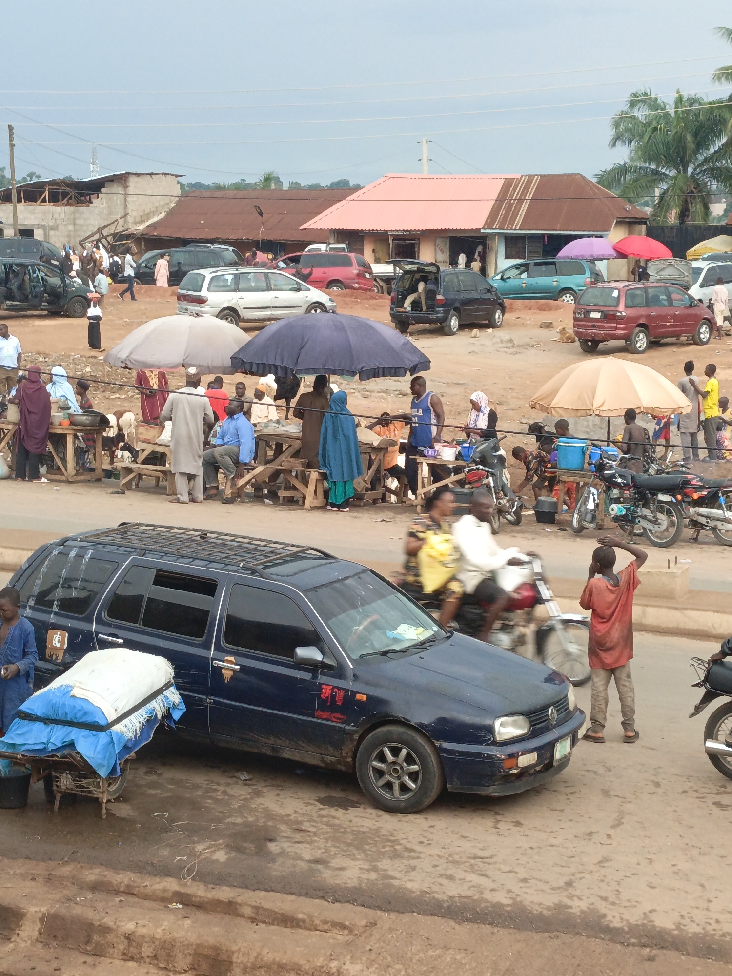 The image depicts a busy roadside scene in Akwanga, Nigeria. A dark blue station wagon car is parked on the left side of the road, next to a cart carrying a large, white bundle covered in blue fabric. Nearby, a person in a blue shirt stands beside the cart. Across the road, a crowded market area is visible, with people gathered around makeshift tables covered by large umbrellas for shade. Some individuals are seated, while others are standing and interacting. Motorbikes are scattered around the market area. In the background, a series of single-story buildings with different colored roofs are visible. The ground is dusty and uneven. Motorbikes speed along the road, past pedestrians.