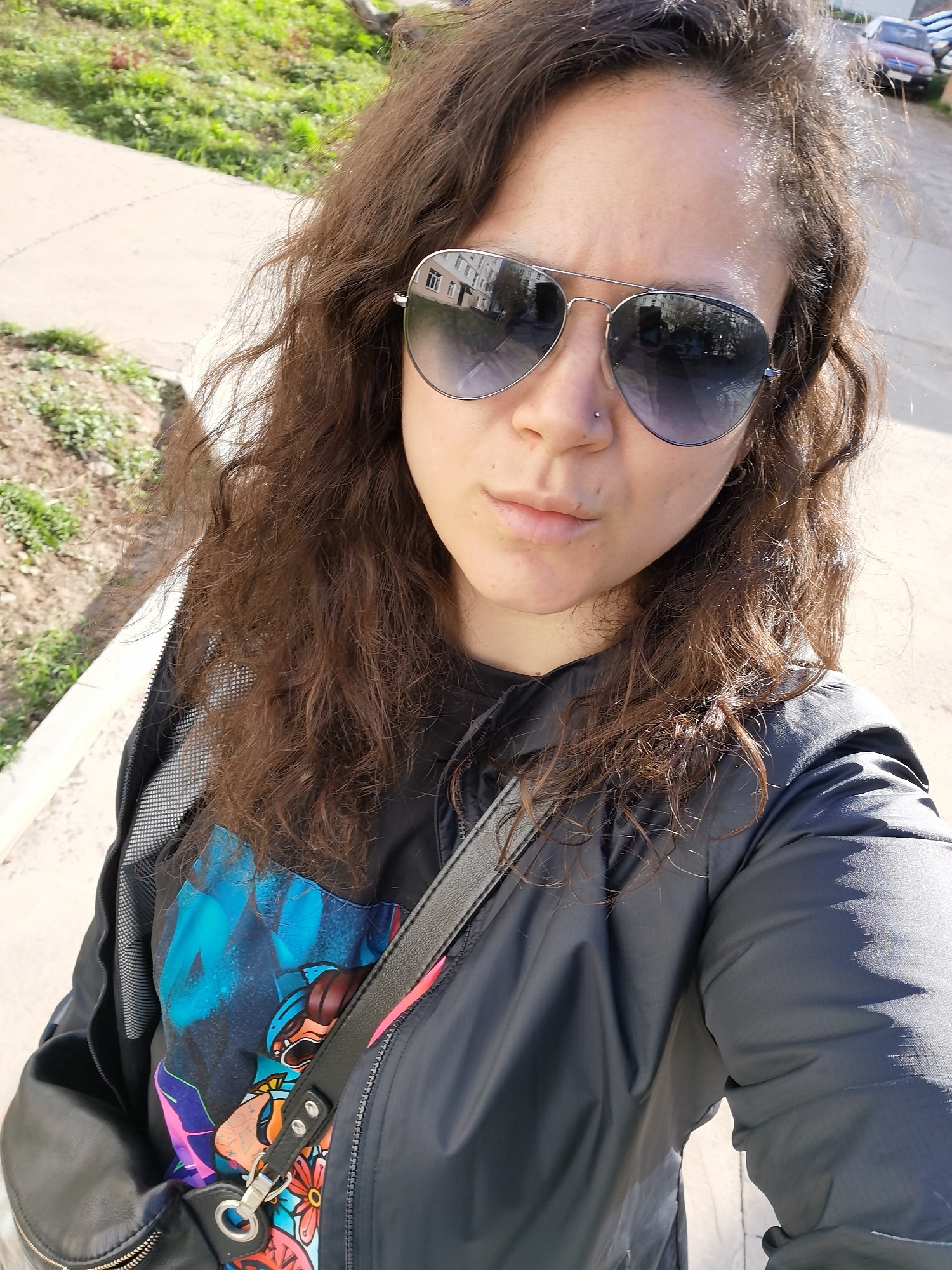 A woman with brown, curly hair is standing outside on a sunny day. She is wearing aviator sunglasses, a black jacket, and a t-shirt with a colorful print. A black shoulder bag is slung across her body. The location appears to be in Chaikovsky, Russia, as the reflection in her sunglasses shows a building typical of the city's architecture. The background includes a sidewalk, grass, and parked cars.