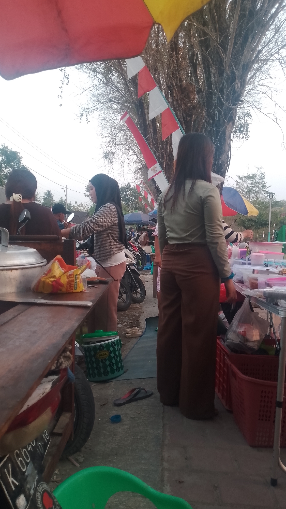 In Karanglangu, Indonesia, an outdoor market scene unfolds under a cloudy sky. A red and yellow umbrella provides shade, while Indonesian flags, red and white, are strung overhead. At a food stall, a woman in a hijab and patterned shirt serves customers. To her right, another woman in a green shirt and brown pants stands. In the foreground, a wooden cart displays a large metal pot, a bag of snacks, and other items. A green plastic bin and red baskets are visible on the ground, along with discarded flip-flops and a blue bottle cap. Motorcycles are parked behind the stall, indicating a busy marketplace.