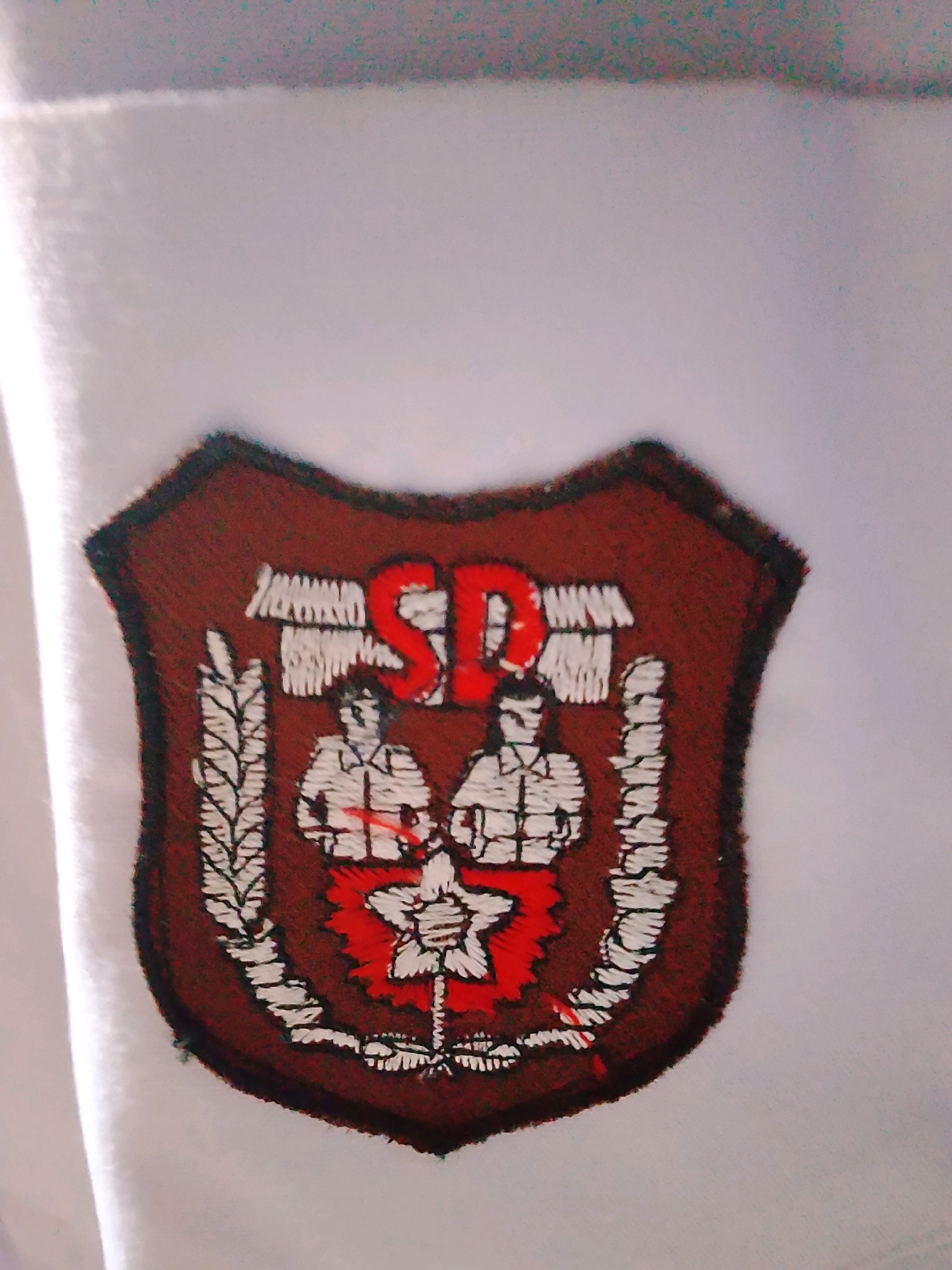 This is a close-up, eye-level, full shot of a patch on a white fabric, likely a uniform or piece of clothing. The patch is shield-shaped with a dark red border and a lighter red interior. In the center of the patch are two figures embroidered in white, appearing to be men in uniform with hats and buttoned shirts. Above them, in large red letters, are the initials "SP". Below the figures is a white star with red detailing. The patch is embellished with white embroidered details resembling wheat stalks on either side. The white fabric appears to have a textured weave. The image is slightly blurry and has some digital noise, particularly in the upper portion, with hints of green and blue pixels. There is no discernible environmental context within the city of Gebang, Indonesia, as the image solely focuses on the patch.