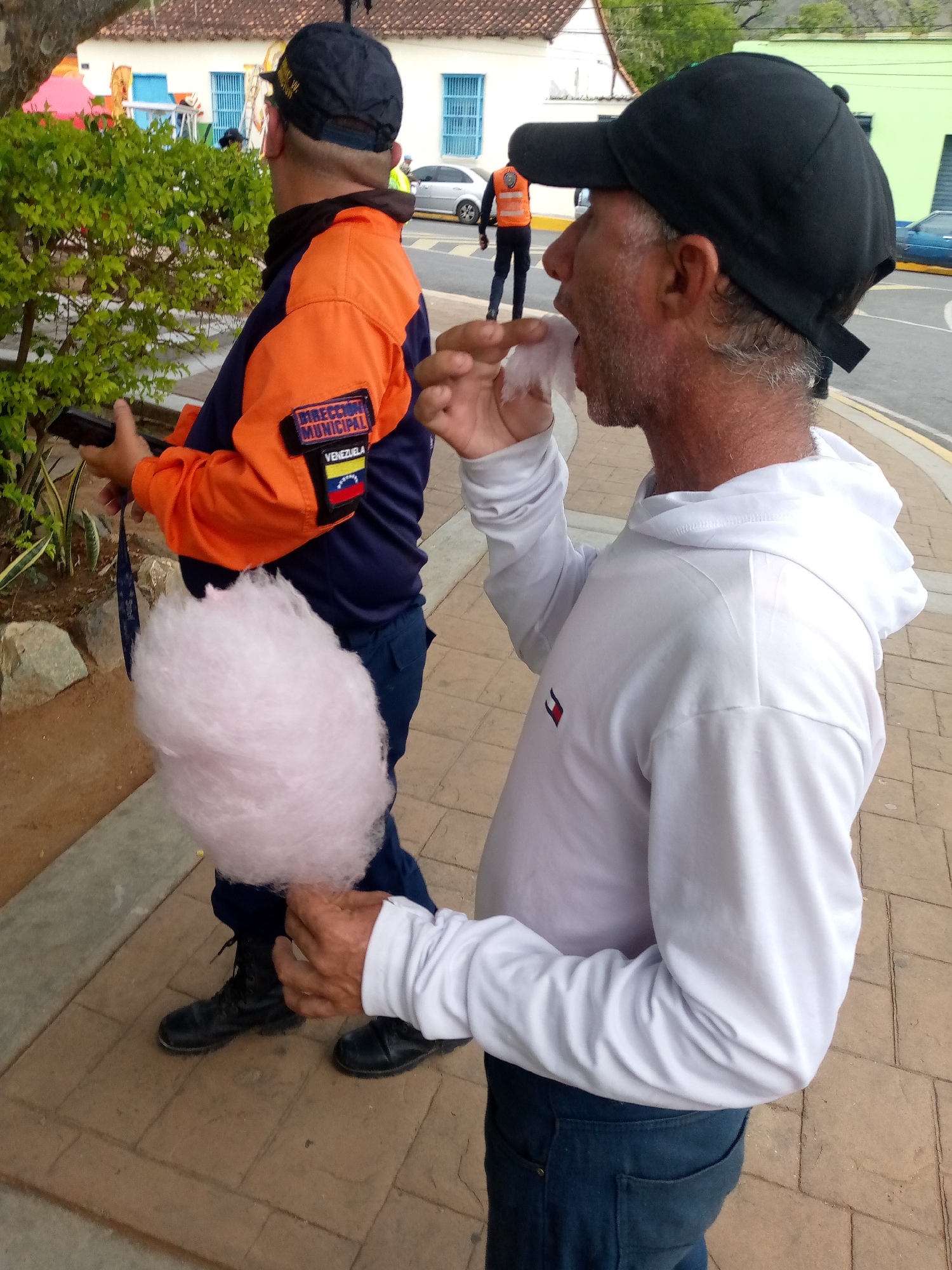 In San Juan Bautista, Venezuela, a man in a white hoodie and dark jeans is eating pink cotton candy on a brick-paved sidewalk. Behind him, another person wearing a navy blue uniform with orange sleeves and a black baseball cap is holding a black object, possibly a phone, and a pink cotton candy. The uniform has a patch that reads "DIRECCION MUNICIPAL VENEZUELA". The background shows a building with white walls and blue windows, some green foliage, and a street with parked cars.