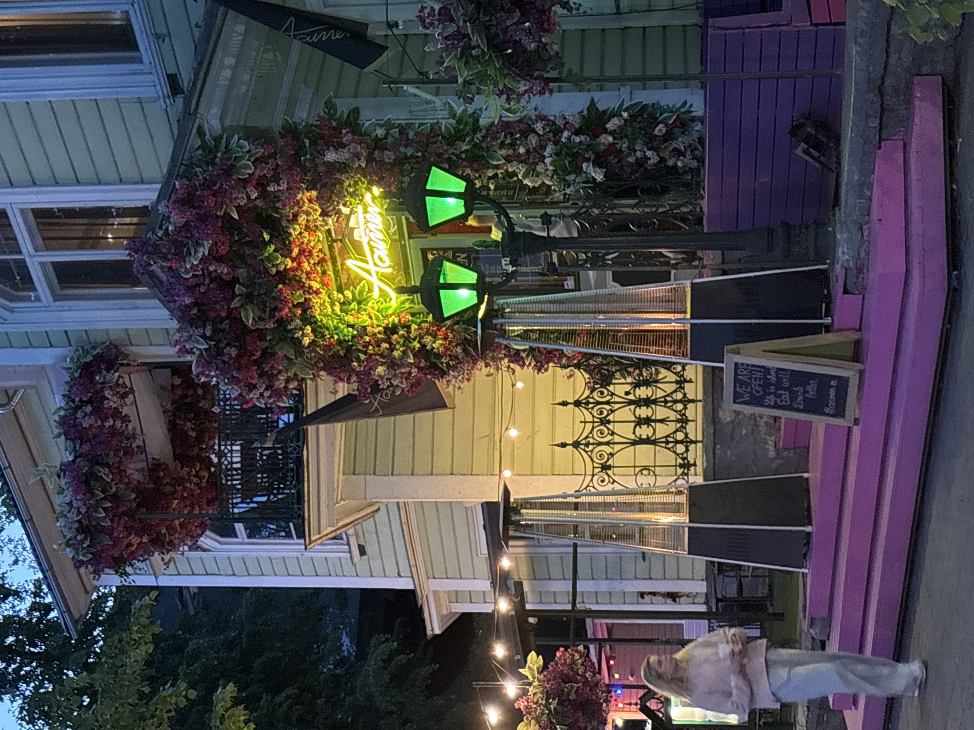 The image depicts an outdoor scene in front of the restaurant "Haamer" located on Lai street in Tallinn Old Town, Estonia, during dusk or early evening. A multi-story, pale green building with white trim features a second-floor balcony and entrance, both heavily adorned with clusters of artificial deep red and purple flowers and green foliage. A prominent bright green neon sign displaying the word "Haamer" is centered above the main entrance, embedded within the floral archway.
In front of the building, a set of outdoor steps and benches are painted a vibrant magenta. Two tall, pyramidal outdoor patio heaters, silver and black, are positioned on these steps, emitting a warm glow from their tops. A black chalkboard sign, standing on the magenta steps to the right, reads "WE ARE OPEN! Eat well, Drink better." in white text. An ornate black metal lamppost stands between the two heaters, featuring two lanterns emitting a bright green light.
To the far left, a woman with long blonde hair, wearing a light pink sweater and light-colored wide-leg pants, walks away from the camera while holding a white cup. String lights are visible hanging across an outdoor area to the left. The sky overhead is a dark blue, indicating the time of day, with ambient light still present. A wrought-iron fence or railing is visible near the right side of the building, above the magenta benches.