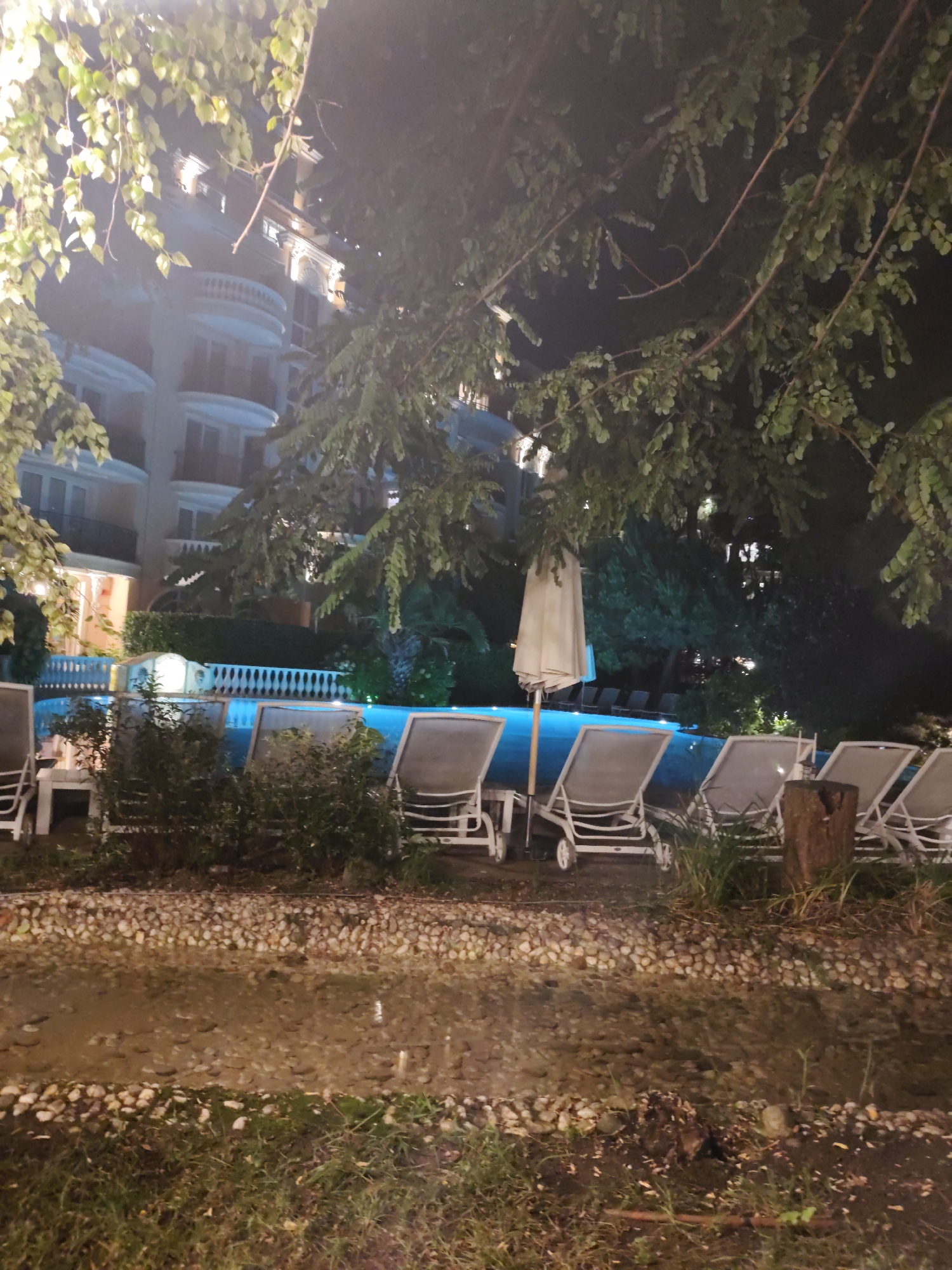 The image depicts a nighttime view of a hotel swimming pool area in Nesebar, Bulgaria. The scene is illuminated by artificial lights, creating a somewhat dim and atmospheric setting. In the background, a multi-story hotel building with architectural details like balconies and decorative elements is visible, its windows glowing with light. In the mid-ground, a swimming pool with luminous blue water is the central feature, bordered by a white ornate railing and lush green foliage. Several empty lounge chairs are arranged around the pool, some with parasols providing shade. The foreground shows a pathway or decorative border made of small stones and pebbles, with patches of grass and some fallen leaves. The overall impression is of a serene, albeit empty, resort setting at night.