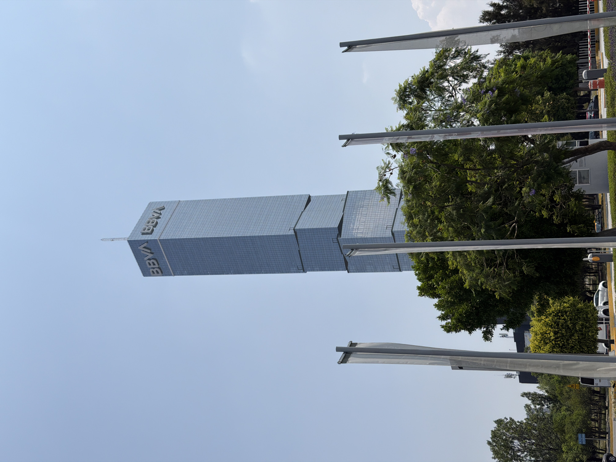 The image shows a tall, modern building in San Bernardino Tlaxcalancingo, Mexico. The building is primarily glass and steel with a stepped design. The BBVA logo is prominently displayed on the upper part of the building. Trees and foliage partially obscure the base of the building. Light poles are present in the foreground. The sky is light blue.