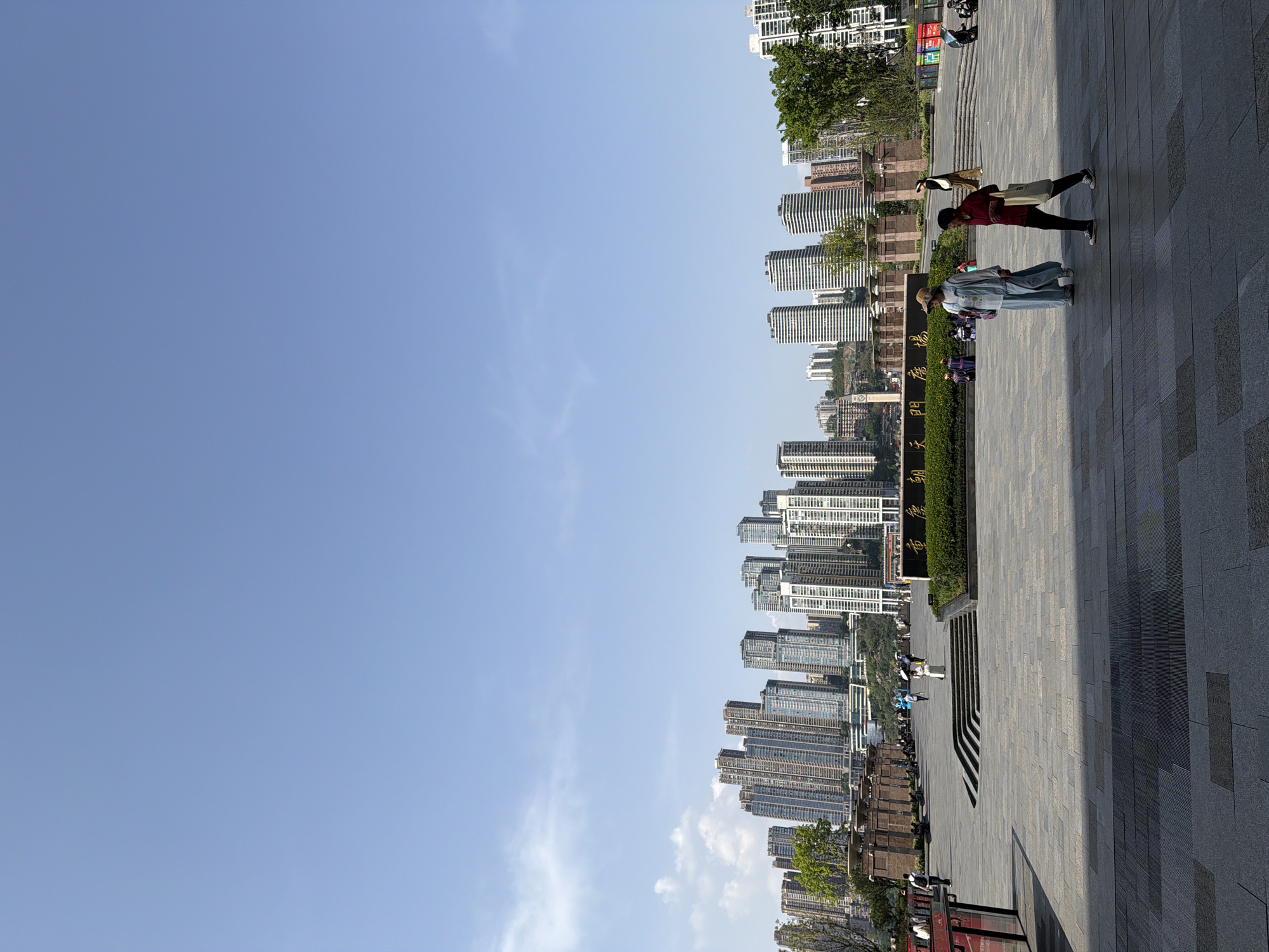 The image depicts an outdoor scene in Yuzhong District, Chongqing, China. The sky is clear and blue. High-rise buildings line the horizon. Pedestrians walk on a paved area, including two individuals walking together. There are architectural elements visible near the bottom of the image.