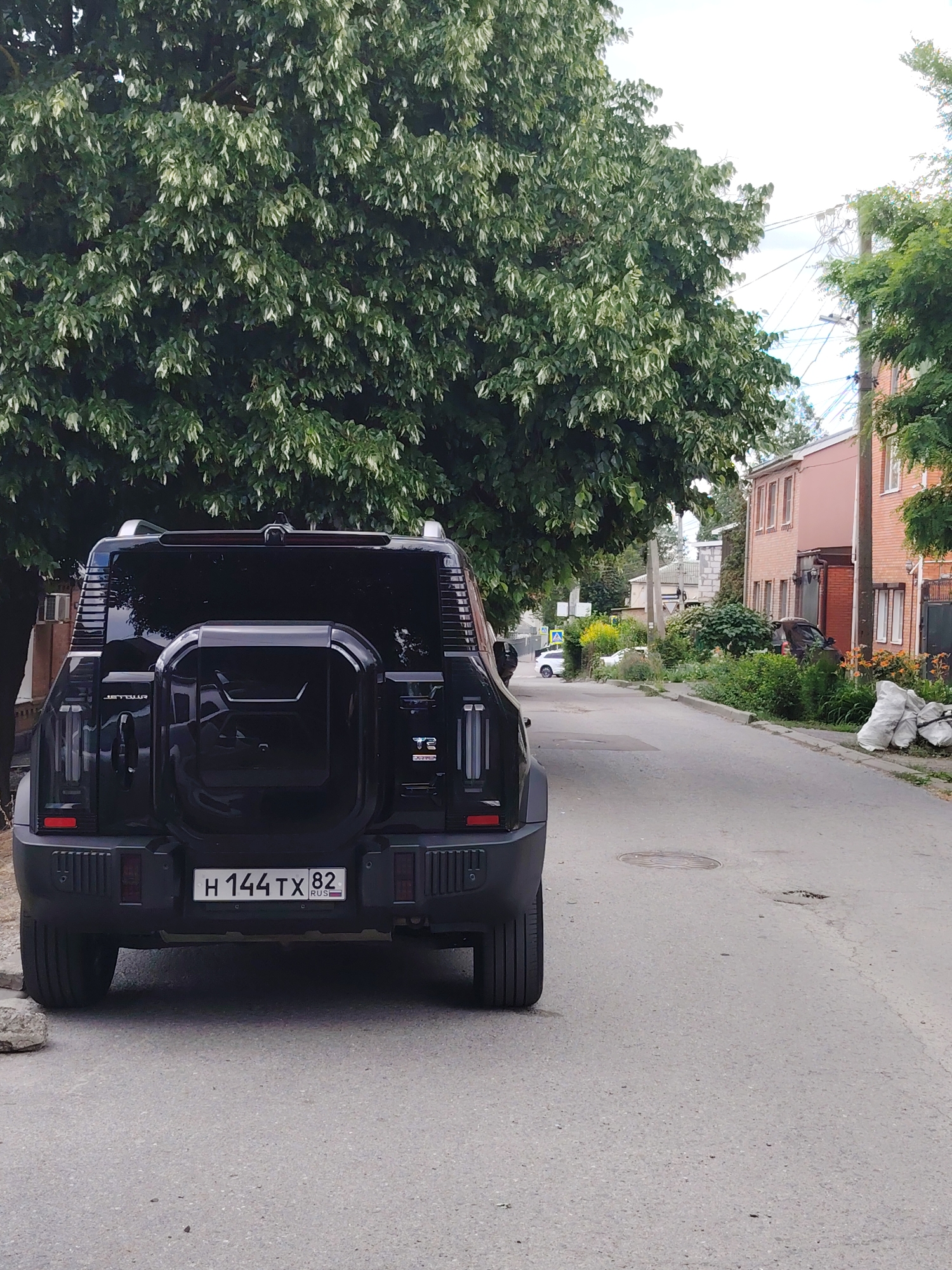 The image depicts the rear view of a black SUV parked on a paved street. The vehicle has a spare tire mounted on its rear and a license plate that reads "H 144TX 82 RUSSIA". A large tree with dense green foliage is visible behind the car, partially obscuring it. In the background, there are buildings, a sidewalk, and more trees. The sky is overcast with some clouds. The environment appears to be a residential or suburban area.