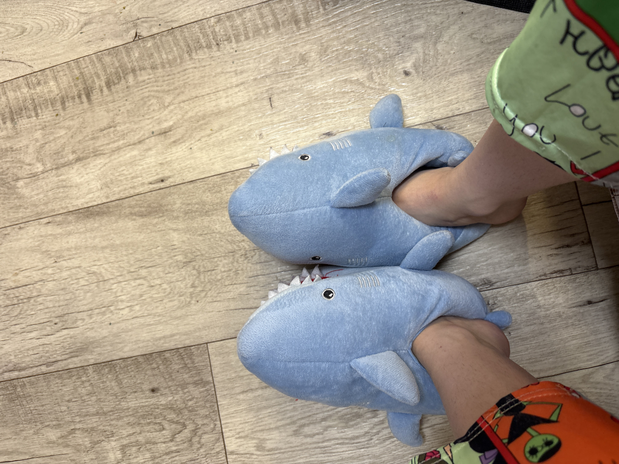 A top-down view shows two feet wearing light blue, plush shark slippers on a light brown, wood-look floor. The left foot is encased in a slipper, with the shark's teeth, eyes, and fins visible. Similarly, the right foot is also in a shark slipper, but the wearer also has on green and red pajama pants with "Love You" written on them. The flooring appears to be laminate or vinyl planks. There is no additional context to suggest a location in the city of Прикамский, Russia.