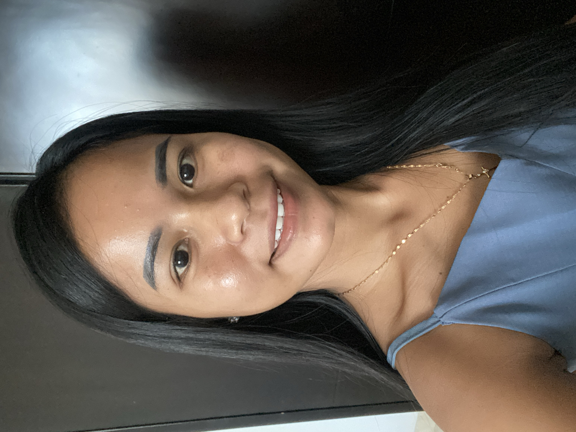 A woman is centered in the frame, facing forward and slightly to her left. She has dark hair, brown eyes, and fair skin. She is wearing a blue top and a delicate gold necklace. The background is nondescript, appearing to be a room interior with dark and light areas, possibly a wall and ceiling. The location is not possible to determine from the image content.